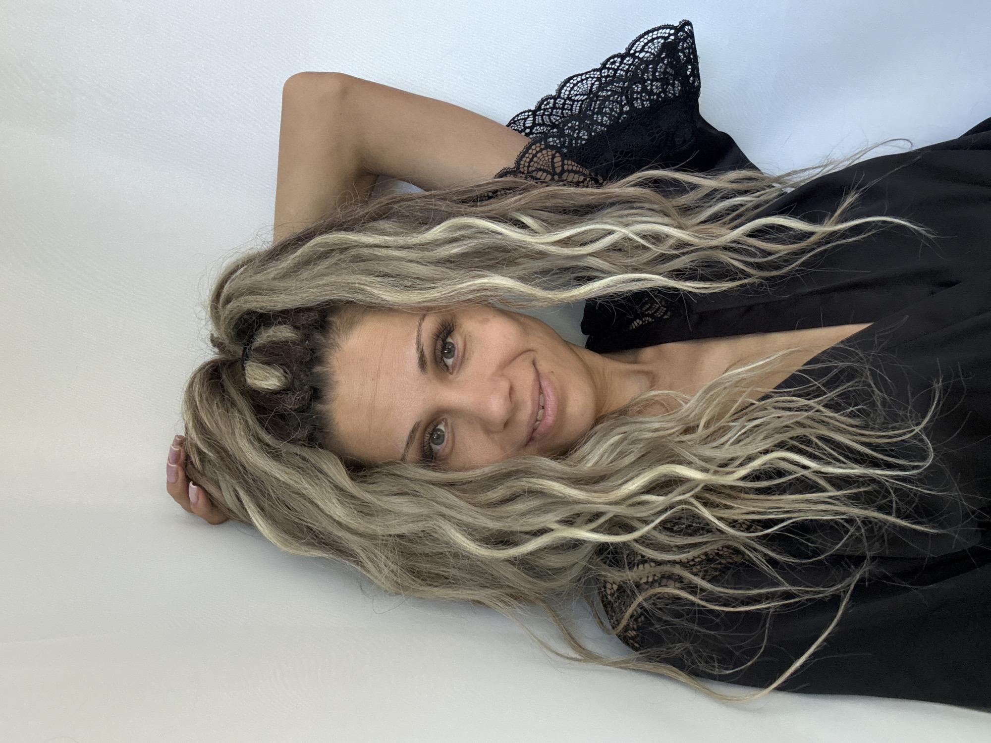 A woman with long, wavy blonde and brown hair tied up in a high ponytail is leaning against a white wall. She is wearing a black robe with lace detailing on the sleeves and neckline. She is looking at the camera with a slight smile. The image is taken from the chest up.