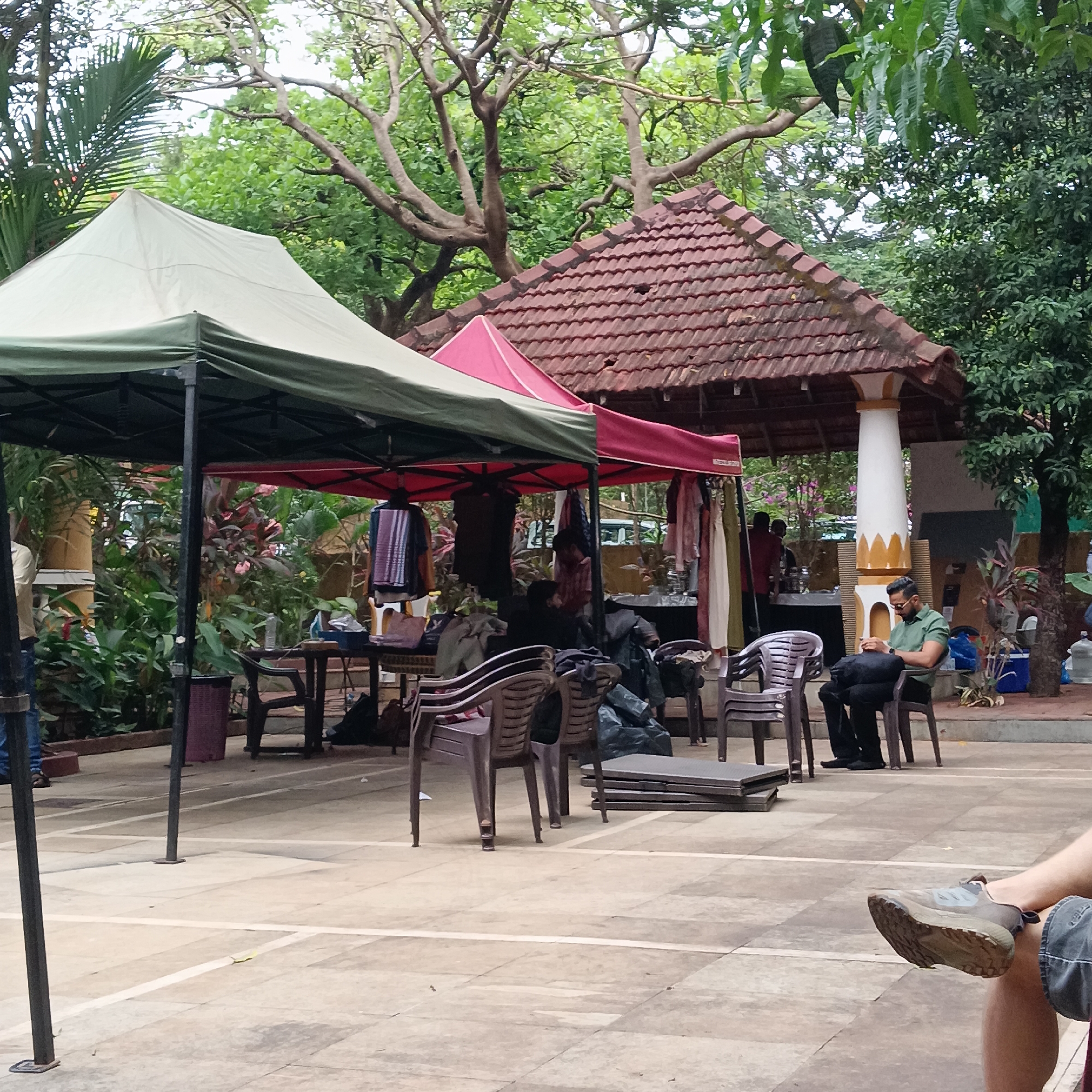 The image shows an outdoor market scene, likely in Anjuna, India. Two tents, one green and one red/green, are set up for vendors. Clothes and bags are displayed under the tents. A man is sitting on a chair looking at a phone. The background includes a traditional Indian structure with a tiled roof supported by pillars. Several plastic chairs are scattered around, and some are stacked. The ground is paved with square tiles. A partial view of a person's leg and shoe is visible in the foreground. The area is surrounded by lush green foliage, typical of a tropical environment.