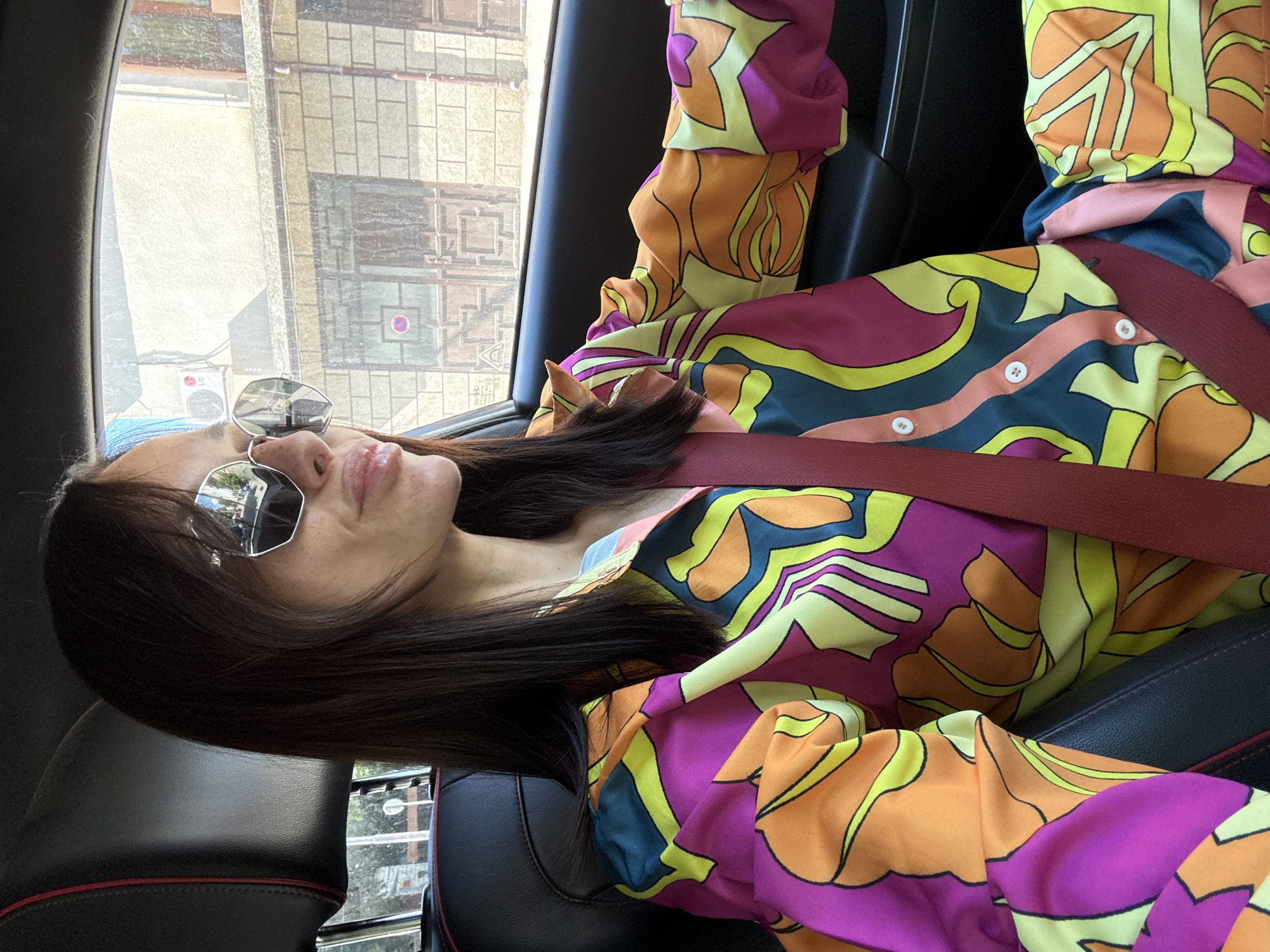 A woman is seated in the driver's seat of a car, wearing sunglasses and a vibrant, multi-colored patterned shirt. The car's interior is visible, with black leather seats and a seatbelt fastened across her chest. Outside the car window, buildings with brick facades and windows can be seen, suggesting an urban environment. The scene is illuminated by daylight. The location is identified as Pleven, Bulgaria.
