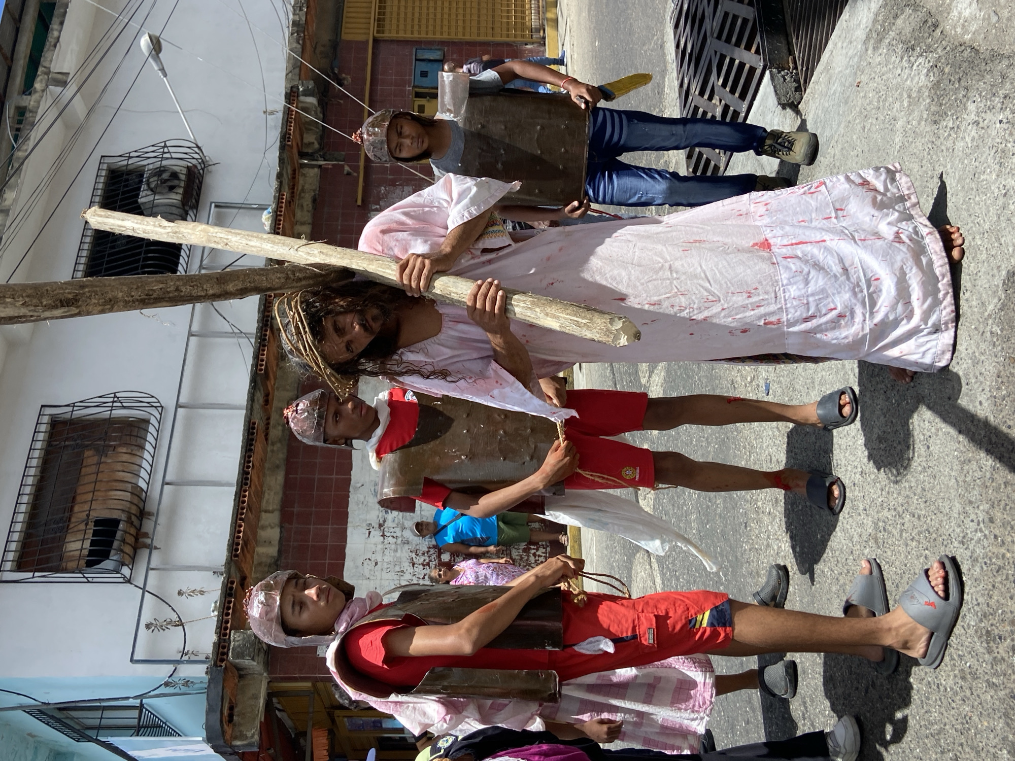 A group of people enact a religious procession on a street in San José de Barlovento, Venezuela. A man portraying Jesus Christ is carrying a wooden cross, wearing a white robe stained with red markings simulating blood and a crown of thorns. He is barefoot. Two people dressed as Roman soldiers flank him, wearing cardboard armor and helmets. A third person follows behind, also in costume. The street is lined with buildings, and a sewer grate is visible on the right side of the image.