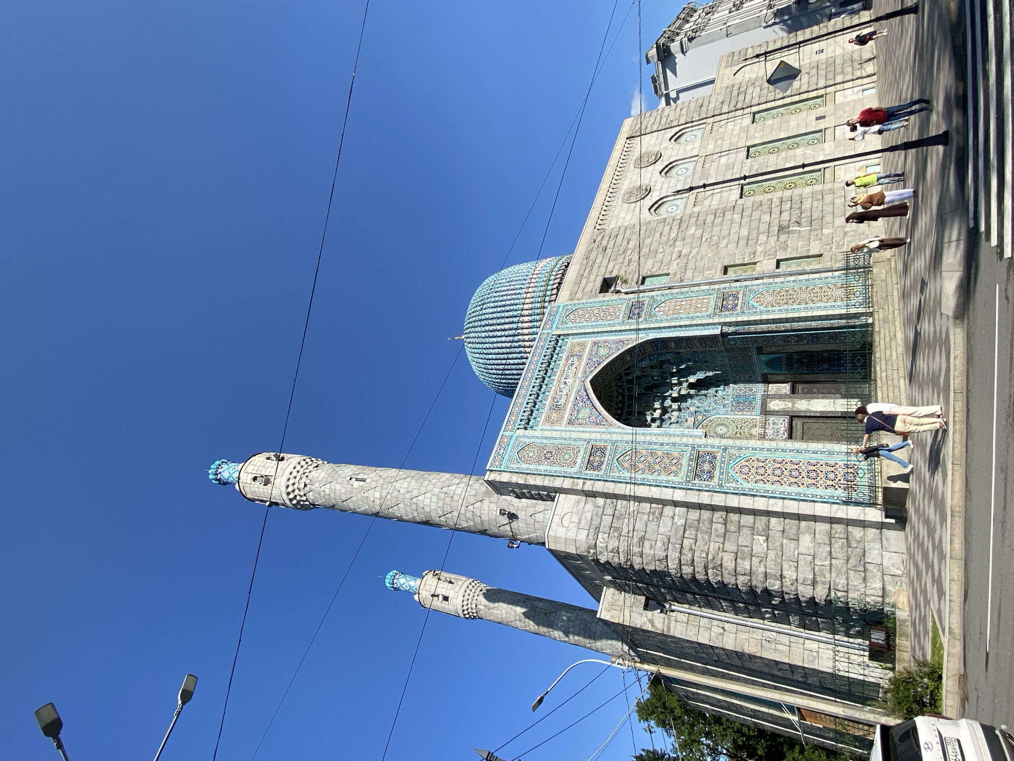 This is a ground-level, wide-angle, outdoor photograph of the Saint Petersburg Mosque in Saint Petersburg, Russia. The mosque is a large, stone building with two tall, slender minarets topped with blue decorative elements. The central dome of the mosque is also a prominent feature, adorned with intricate blue tiling. The entrance to the mosque features a grand archway decorated with colorful mosaic tiles. Several people are visible on the sidewalk in front of the mosque, some walking and some standing. A street with a crosswalk is in the foreground, and a white car is parked on the left side of the frame. Power lines are visible crisscrossing the bright blue sky. The architecture suggests a Moorish or Central Asian influence.