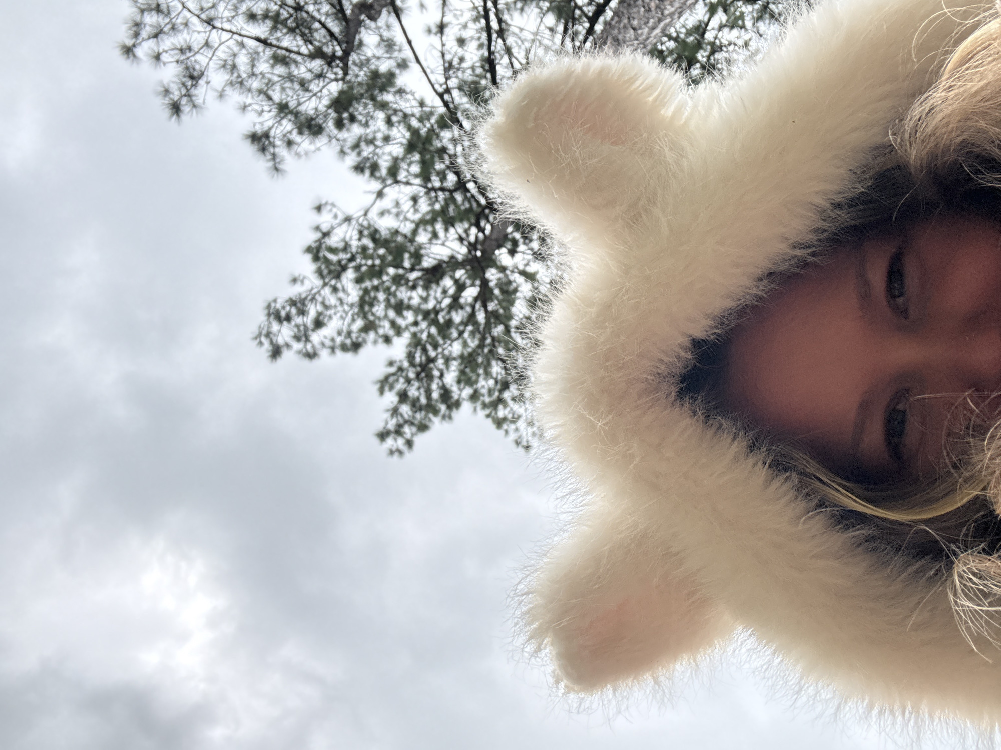 The image shows a close-up of a person wearing a white fuzzy hood with two ear-like protrusions, angled upwards towards a cloudy sky and pine trees. The person's face is partially visible from the nose down, with blondish hair and an eye visible on the right side of the frame. The location is not specified within the image or its context to be in Đạ Cháy, Vietnam.
