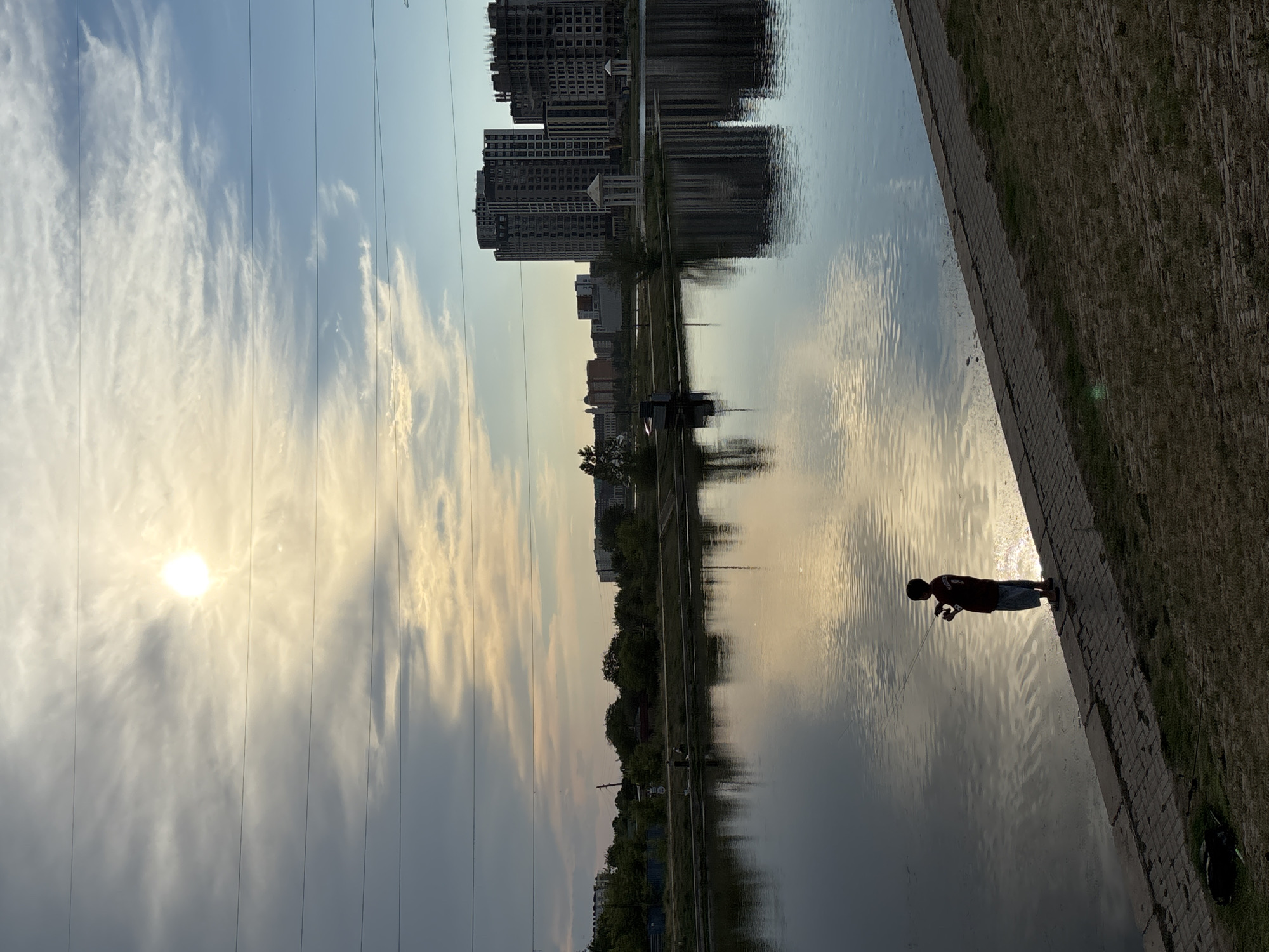 This image depicts a waterside scene with a silhouette of a child fishing in the foreground, located in Astana, Kazakhstan. The child, facing the water, is holding a fishing rod. The body of water, likely a canal or river, reflects the sky and the surrounding cityscape. Across the water, a developed area is visible, featuring modern high-rise buildings under construction and some completed structures, indicative of urban development. Trees and other vegetation line the banks. The sky is partly cloudy with the sun casting a bright glow, suggesting it is either sunrise or sunset. Overhead power lines are visible, a common feature in urban environments. The ground in the foreground is a mix of paved walkway and grassy embankment.