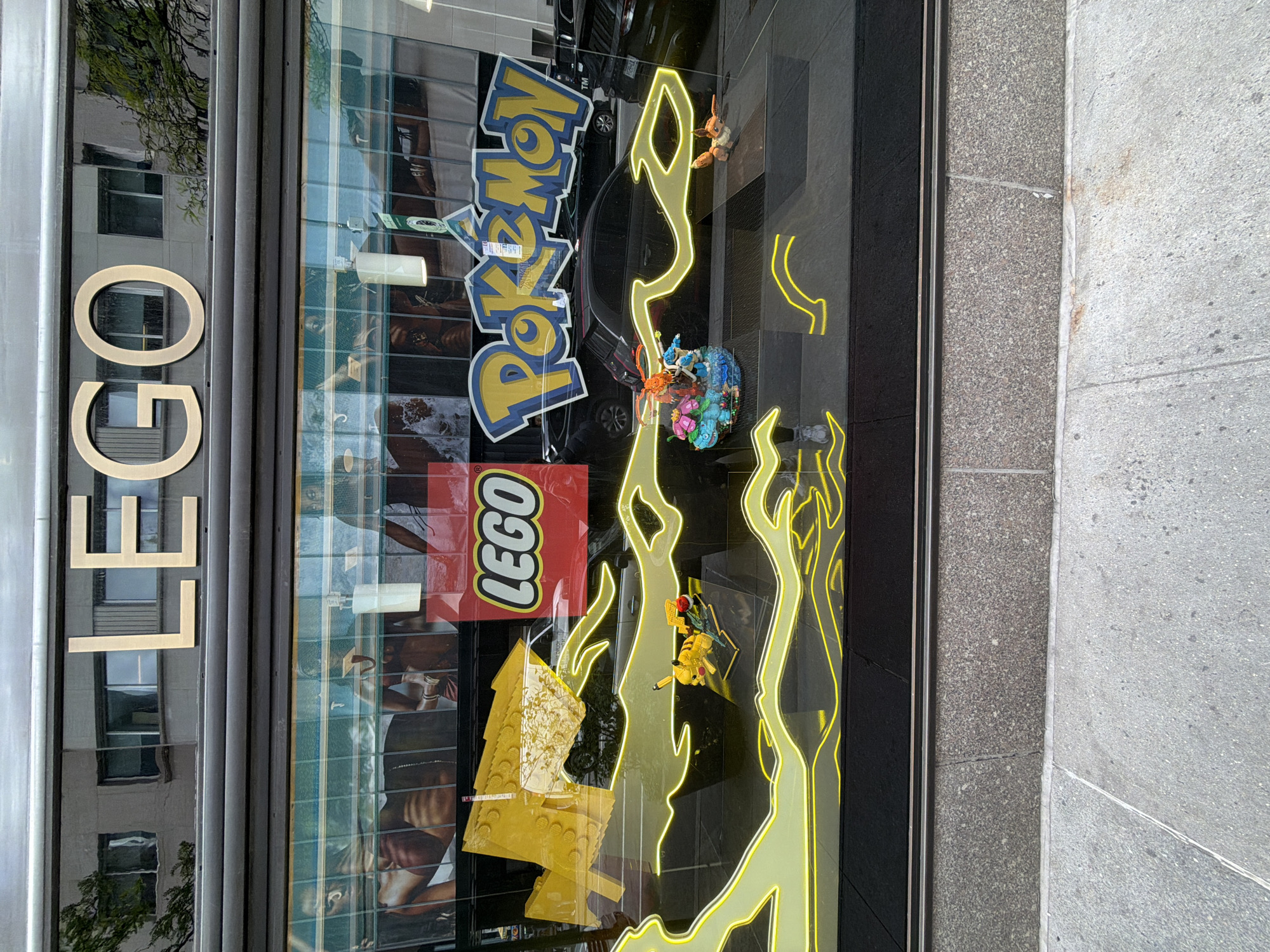 The image shows a retail storefront in New York City, United States of America. The sign on the side of the building reads "LEGO" vertically. Inside the window, there's a red LEGO logo and a Pokémon logo. Yellow neon lights shaped like lightning bolts are also visible. A LEGO Pikachu figure and another LEGO model resembling a Pokémon character are displayed within the window alongside other LEGO displays. Reflections of buildings and a car are seen in the glass. The storefront is situated on a street with a gray concrete sidewalk.