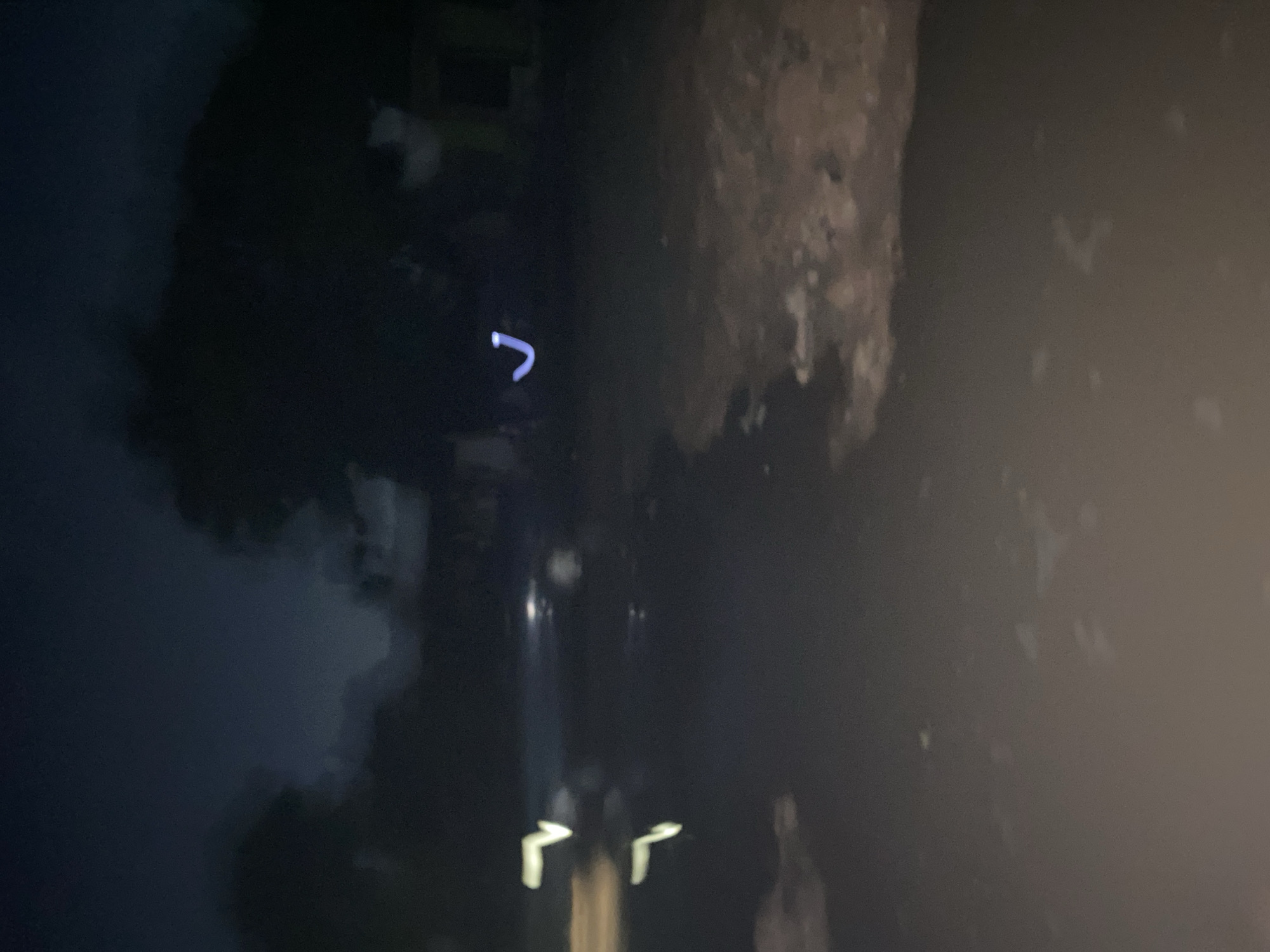 A dark, blurred image depicts a nighttime scene. Two illuminated "L" shapes are visible near the bottom center of the image, likely reflecting off a surface. A blurry, light-colored "hook" shape is positioned in the upper left quadrant. To the right, a large, rough-textured mass is visible. The environment appears indistinct and obscured. The exact location is unknown.