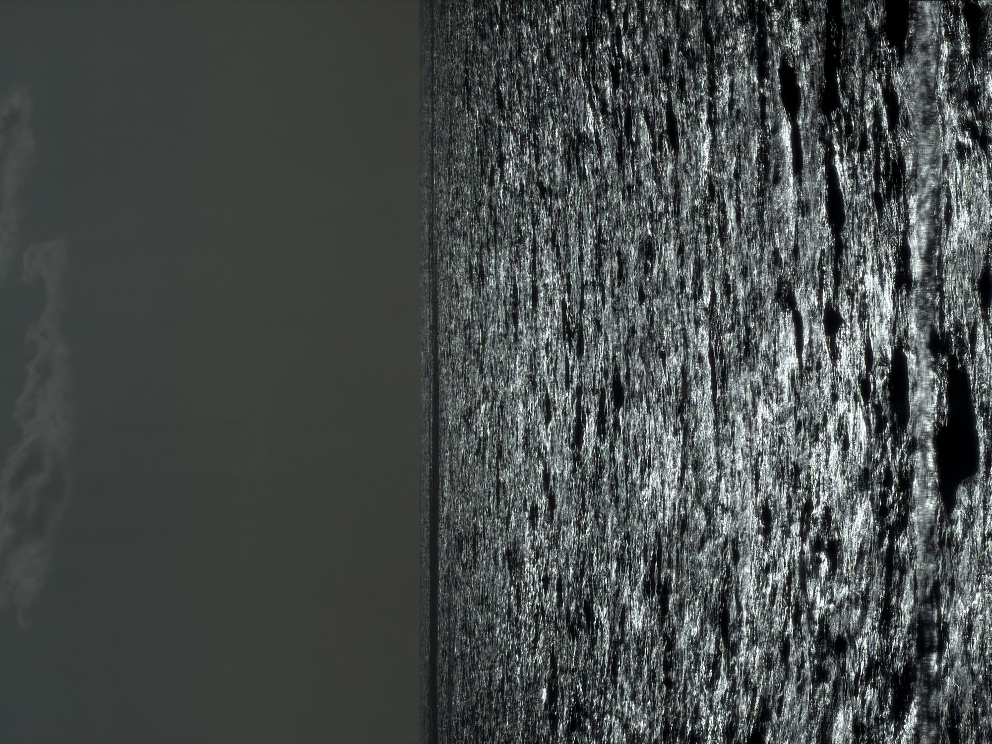 The image depicts a dark, overcast scene. The left side of the image is predominantly a uniform dark gray, possibly representing a cloudy sky. Faint cloud formations are visible. The right side displays a highly textured surface with strong highlights, likely water with strong sunlight reflections. The scene appears to be water meeting the sky on a dark day. Location is unknown.