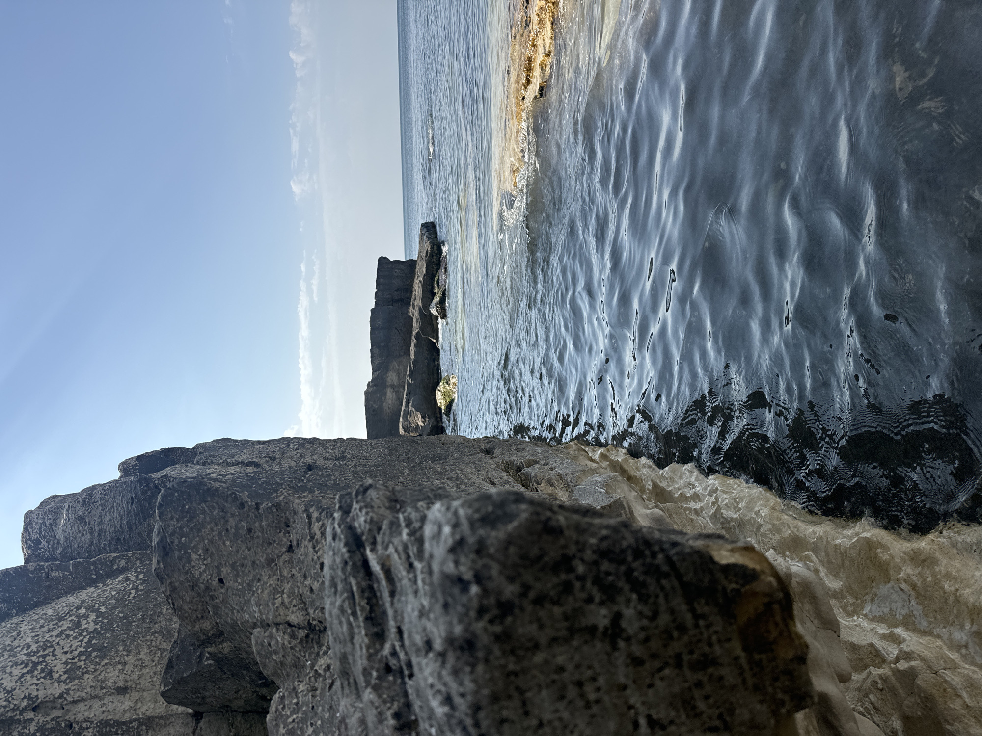 This is a close-up, eye-level, outdoor, daytime, vertical shot of the coastline near Aktau, Kazakhstan. In the foreground on the left, a large, rough, grey rock formation juts out. The rock face is textured and shows layers of sediment. The bottom portion of the rock formation is lighter, appearing wet and possibly encrusted with minerals. To the right of the rock, dark blue water ripples gently, reflecting the sky. The water's surface is disturbed by small waves and shows darker and lighter patches. Further in the background, on the right side of the image, a section of rocky coastline is visible, with flatter rock formations extending into the sea. The water there appears calmer and lighter in color, with some areas of yellowish-brown discoloration near the shore. Beyond that, the sea stretches to the horizon, meeting a clear, pale blue sky with a few wispy white clouds. The overall scene depicts a rugged coastal landscape under a bright sky.
