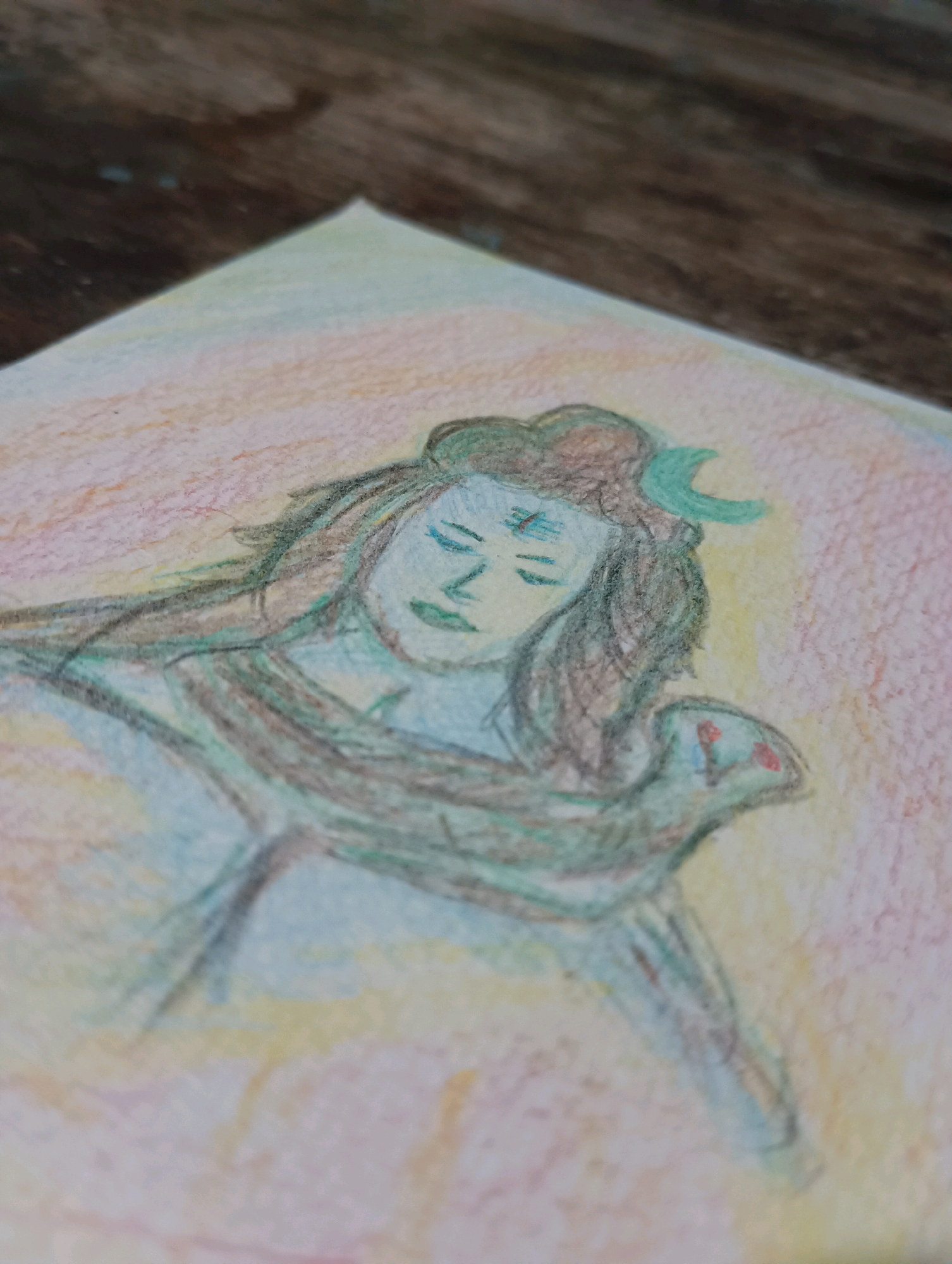 A watercolor painting depicts a figure with serene closed eyes and a crescent moon on its head, reminiscent of the Hindu deity Shiva. The figure has long, dark hair and is adorned with a snake around its neck. The background is a soft blend of pink, yellow, and pale blue, suggesting a tranquil atmosphere. The painting is rendered on textured paper and is partially visible on a wooden surface. There is no information in the image to determine its location in Dakhin Parsa, India.