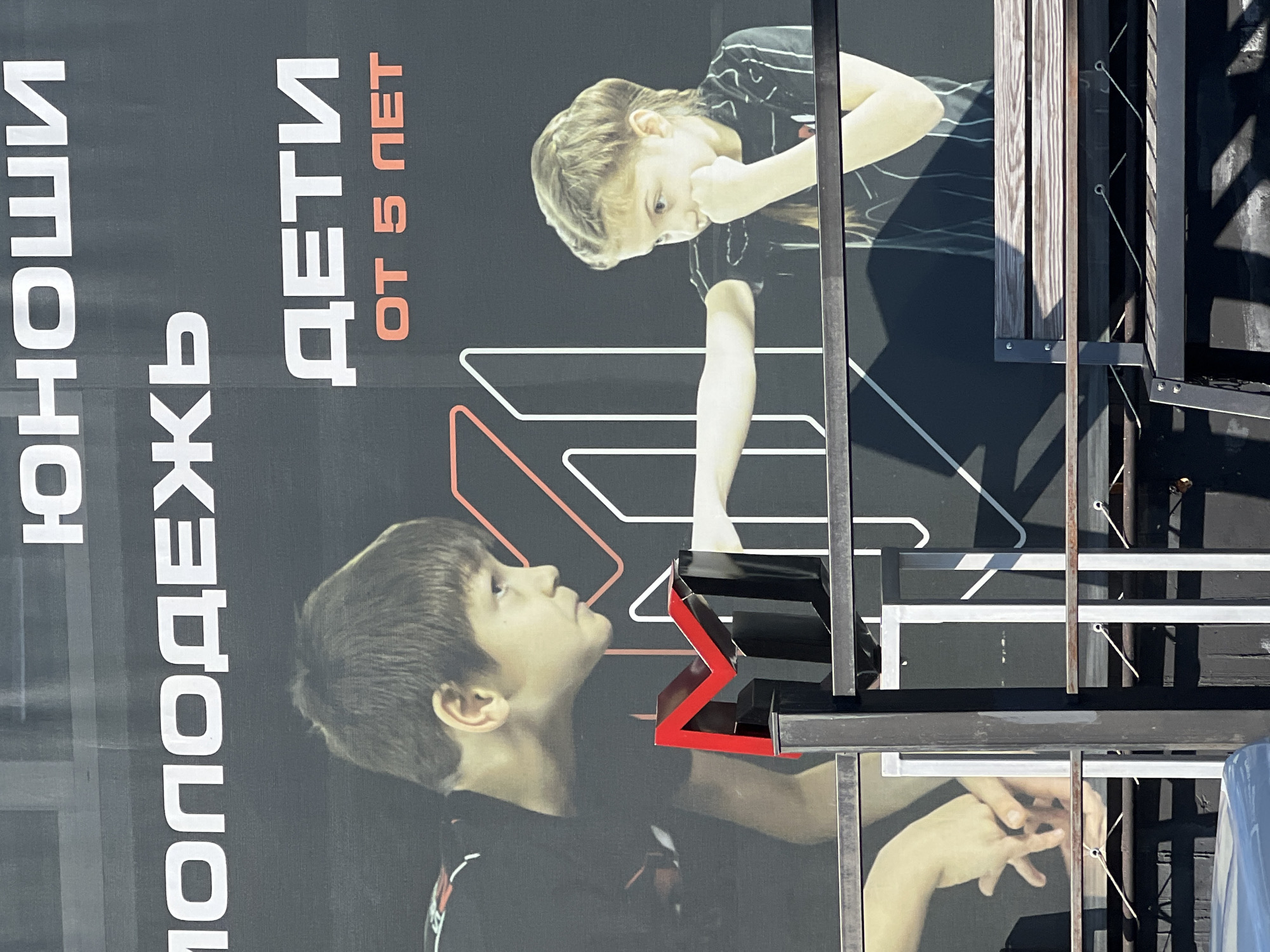 The image shows a billboard advertising a children's sports program, likely martial arts or boxing, in Sochi, Russia. The billboard features large white Russian text that translates to "Youth" and "Children from 5 years old." Two children are depicted: a boy on the left and a girl on the right, both in profile. The boy is shown with his arms bent and hands clasped near his chest, while the girl is in a defensive stance, with one arm extended and the other held near her chin. The children are placed against a black background with stylized red and white lines, possibly representing movement or a sports arena. In the foreground, a metal bench is visible, suggesting this billboard is displayed outdoors, possibly at a sports facility or public space in Sochi. The lighting indicates it is daytime.