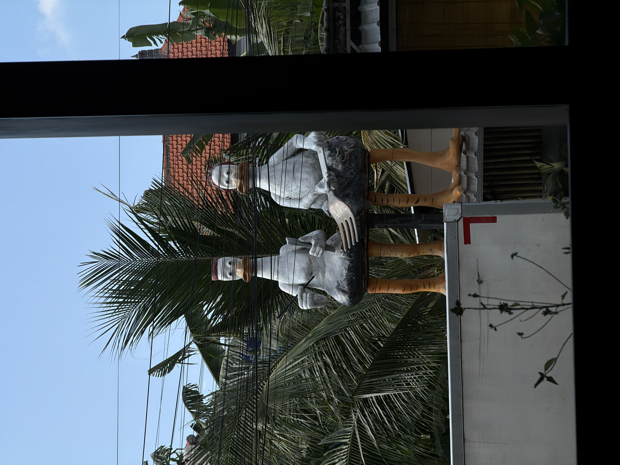 Two stylized statues depicting anthropomorphic birds stand side-by-side, each holding a large fork and knife. The figures are primarily white with black torsos and orange striped legs and feet. One statue has a red cap, while the other has a white cap. Palm trees fill the background. The statues are positioned atop a white vehicle, which has a red stripe on the side. The image is taken through a window, with the window frame partially visible at the top and bottom. The location is likely in Kuta, Indonesia, suggested by the tropical vegetation and the style of the statues.