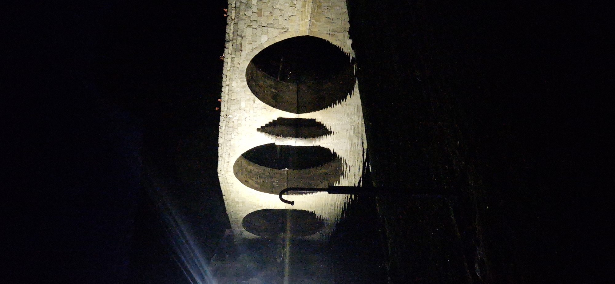 A stone bridge with multiple arches is illuminated at night over a body of water. The bridge, constructed from rough-hewn stones, has a solid parapet along its top. Several figures are visible standing on the bridge's walkway. The water below reflects the illuminated arches, creating a symmetrical effect. In the foreground, a dark, silhouetted object resembling a faucet or fountain fixture emerges from the water. The surrounding environment is dark, suggesting nighttime. This scene is of the Puente Romano, a Roman bridge located in Navaluenga, Spain.