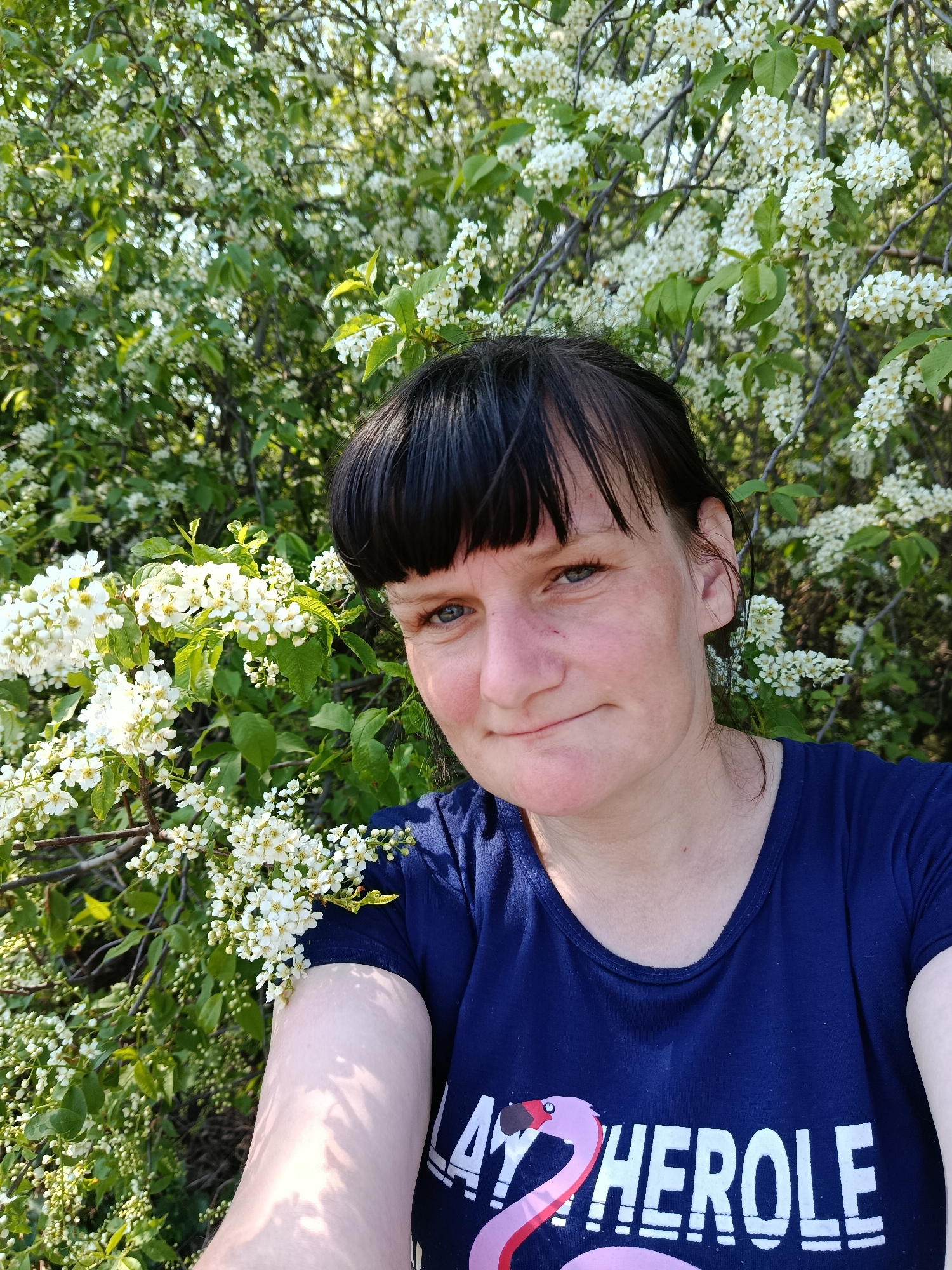 A medium shot shows a woman with short dark hair, blue eyes, and fair skin posing in front of a flowering tree or bush. The woman wears a dark blue t-shirt with a graphic of a flamingo and the word "HEROLE" in white letters. The tree or bush behind her is covered in clusters of small white flowers and green leaves. The background is a blur of greenery. The photo appears to be taken outdoors in daylight. The location is Фоки, Russia.