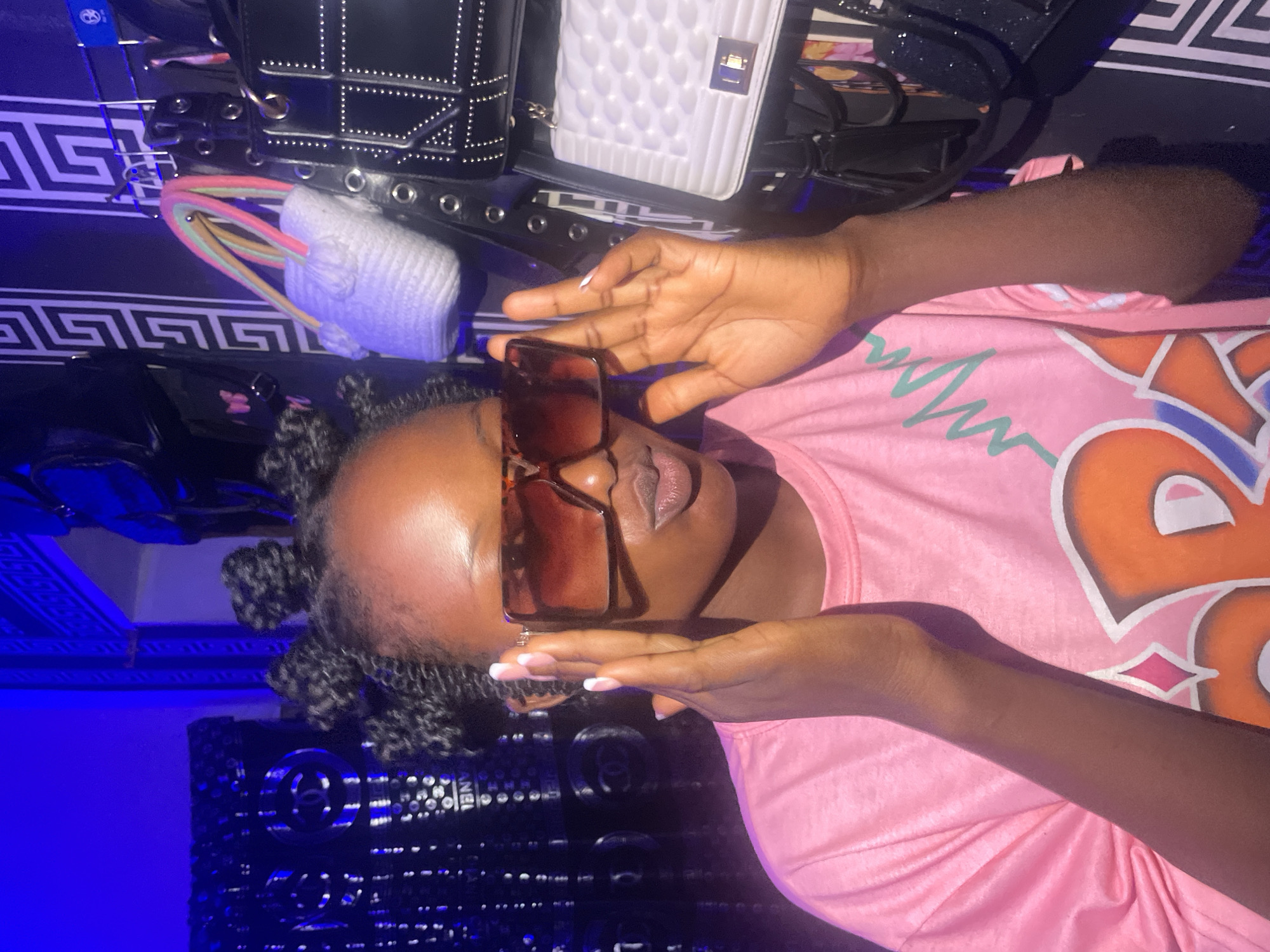 The image shows a dark-skinned person wearing a pink shirt and brown sunglasses. The person is lying down, with their hands near their face, as if posing for a photo. The background includes several bags, a shelf with items on it, and a wall with a Greek key pattern. A blue light is present, which could be from a light fixture or reflection. There is not enough information to determine the exact location in Lokoja, Nigeria.