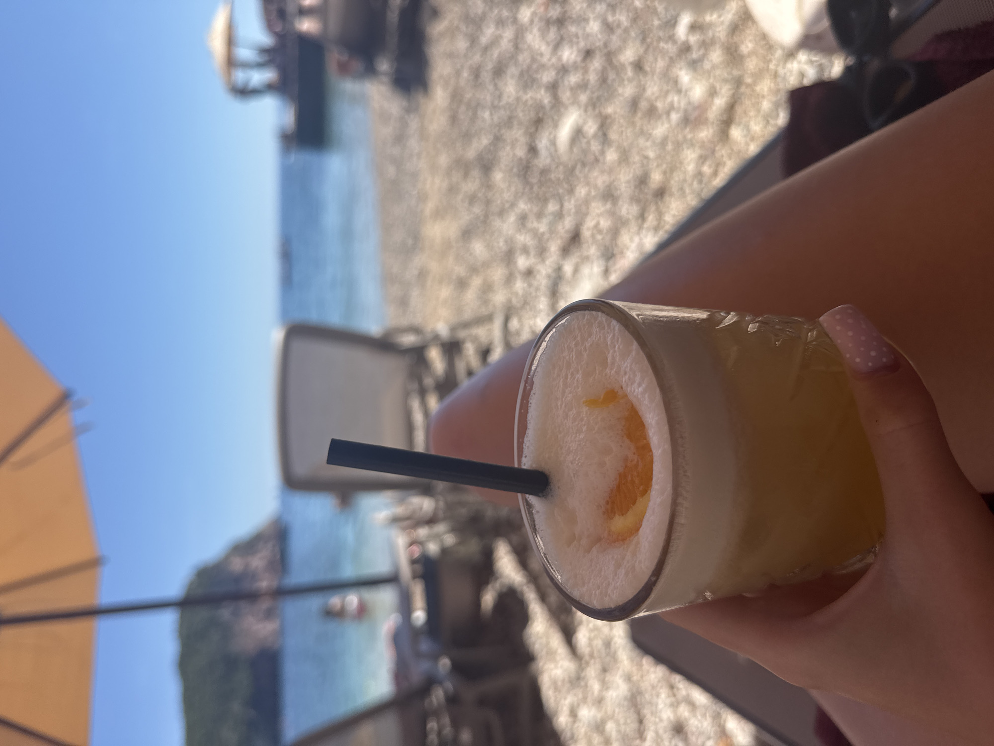 An overhead perspective shot captures a hand holding a cocktail on a pebbly beach. The drink, topped with a frothy layer and garnished with an orange slice, features a black straw. In the background, the turquoise ocean meets a clear blue sky, with a distant green, rocky island on the left. Beach chairs and umbrellas are visible, with people dotted along the shore, suggesting a leisurely day by the sea. The sun is shining brightly, illuminating the scene.