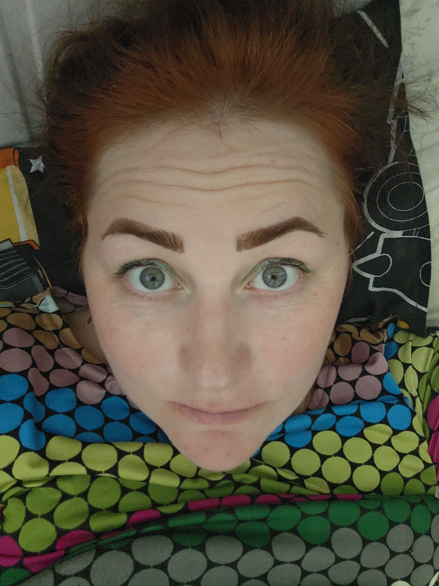 A close-up, high-angle shot shows a woman with red hair, light skin, and freckles lying on what appears to be a bed. Her gaze is directed upwards towards the camera. She has light blue or gray eyes with green eyeliner. Her eyebrows are brown and shaped. She is wearing a multi-colored shirt or dress with a polka-dot pattern of blue, pink, green, and gray. The background includes a black pillow with a white geometric design and a yellow pillow with a star. The location is not specified.