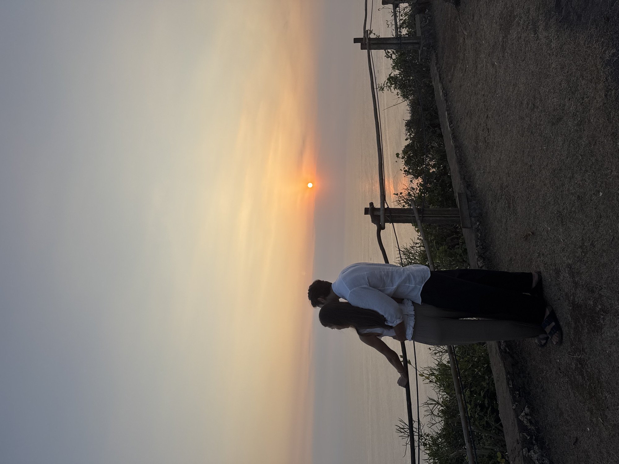 A couple stands embracing on a cliff overlooking the ocean at sunset. The woman is on the left, wearing a white tank top and tan pants. The man is on the right, wearing a white button-down shirt and black pants. They are both facing the ocean, watching the sun dip below the horizon. The sky is a gradient of orange, pink, and blue, with the sun a bright orb in the distance. The ocean is calm, with a faint reflection of the sunset on its surface. A wooden railing separates the couple from the edge of the cliff, and lush green foliage grows around the railing. The ground beneath them is covered in dirt and dry grass.