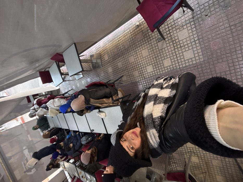 The image depicts a bustling outdoor or semi-enclosed café seating area in Azrou, Morocco, populated by multiple individuals. In the foreground, a woman with long dark hair, wearing a black knit beanie, a black leather-look jacket, and a black and white checkered scarf, takes a selfie, smiling towards the camera with her right arm extended.
Behind her, approximately fifteen to twenty adults and children are seated at various tables. Several rectangular tables with light-colored tops and metal bases are arranged end-to-end, forming long communal rows, while others are positioned individually. The occupants, dressed in winter attire including hooded jackets, scarves, and hats, are seated on red-upholstered chairs with dark frames. They appear to be engaged in a social setting.
The seating area is enclosed by tall, light-colored fabric or tent-like walls, creating a covered space. The floor is composed of a repeating pattern of light and dark brown square tiles. Additional empty tables with red chairs are visible along the right side of the frame. In the upper left background, an opening in the enclosure reveals a glimpse of an outdoor street with distant buildings and a white vehicle.