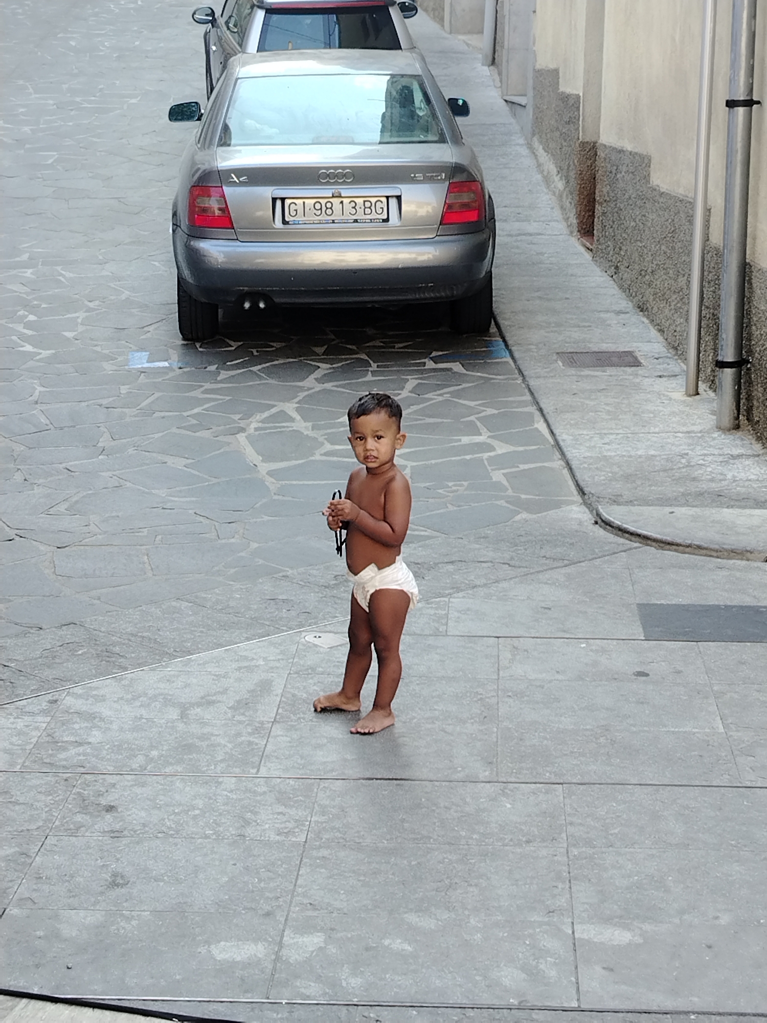 A young child wearing only a diaper stands on a paved street in Hostalric, Spain. The child is facing the camera, looking forward with a neutral expression. The child is holding what appears to be a small black object in their hands. Behind the child, parked on the street, is a silver Audi sedan with a Spanish license plate that reads "GI 9813 BG". The street is paved with irregular grey flagstones. To the right of the child and the car is a textured wall with horizontal bands of dark grey and light grey, and a metal pipe runs vertically along the wall. The scene appears to be an outdoor urban environment.