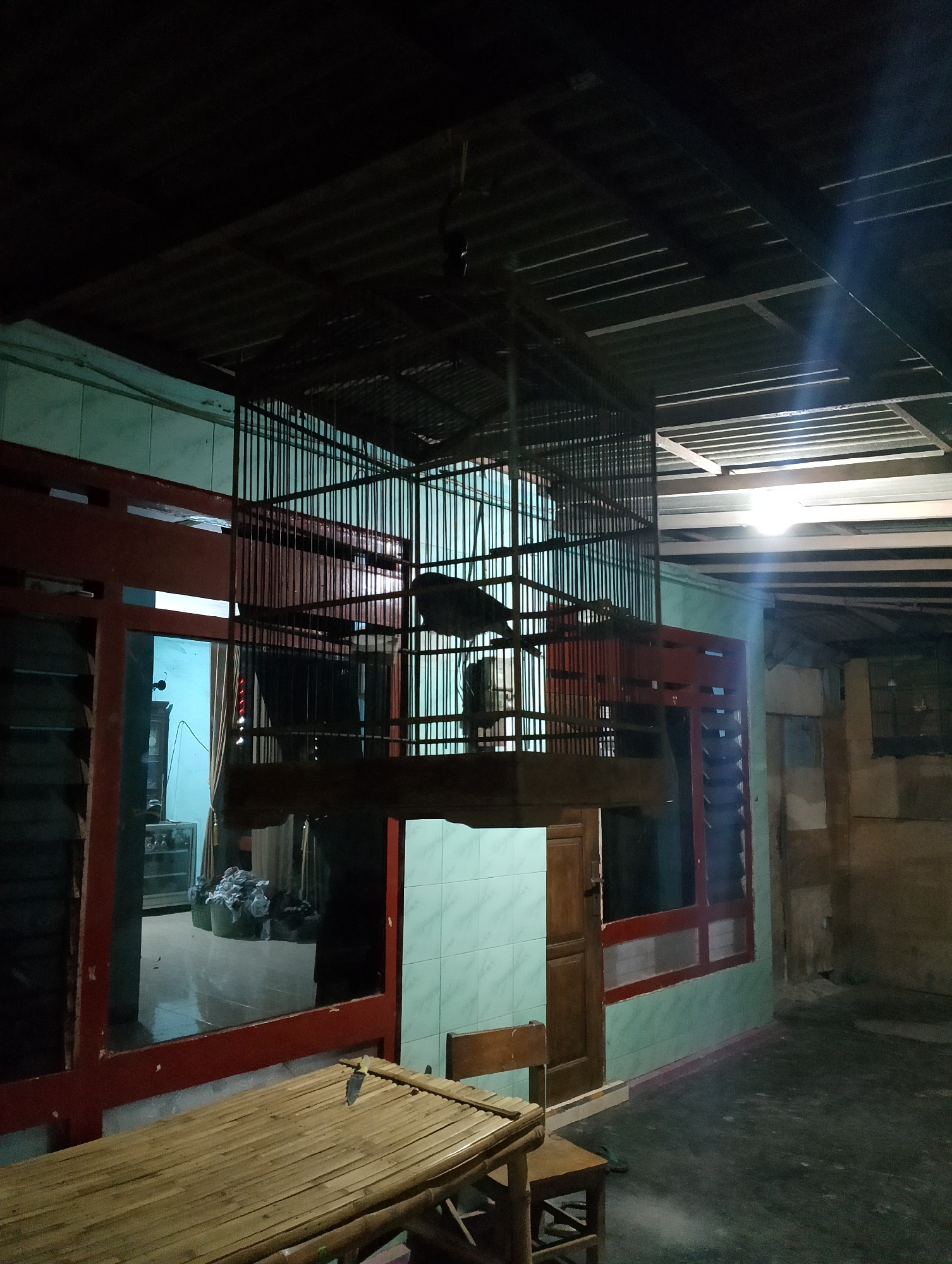 A wide-angle, low-light, exterior shot captures an outdoor covered area, possibly a porch or workshop, attached to a dwelling. The scene is located in Rembang, Indonesia. In the foreground, a low bamboo table with two wooden chairs sits on a dark, dusty concrete floor. A knife rests on the table. Behind the table, a large, dark-colored cage, likely for birds, hangs from the ceiling. The cage contains at least two birds. The dwelling's wall features a light green tiled section and red-trimmed windows. A closed wooden door is visible. The background reveals more of the dwelling, including a reflection in a window showing a room with baskets of laundry and furniture. The ceiling is corrugated metal. The overall impression is one of a functional, utilitarian outdoor space at night, illuminated by a single bright light source creating strong shadows.