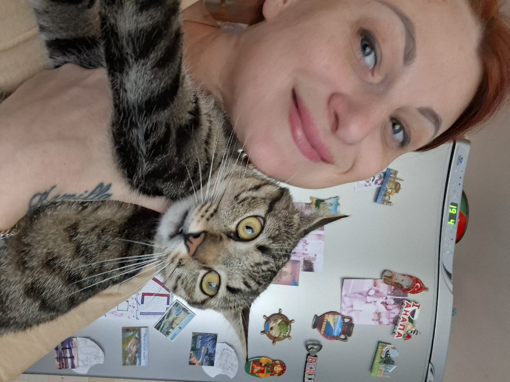 A woman with reddish-brown hair and light-colored skin, wearing a light-colored top, is holding a tabby cat in front of a refrigerator. The woman is smiling and looking to her left. The cat has yellow eyes and is looking forward. The refrigerator is covered with magnets and what appear to be photos or postcards. One magnet reads "I АНАПА" with a Russian flag. Another magnet is a Matryoshka doll. The background appears to be an indoor setting, possibly a kitchen. There is no discernible information about the city of Myadzyel, Belarus in the image.