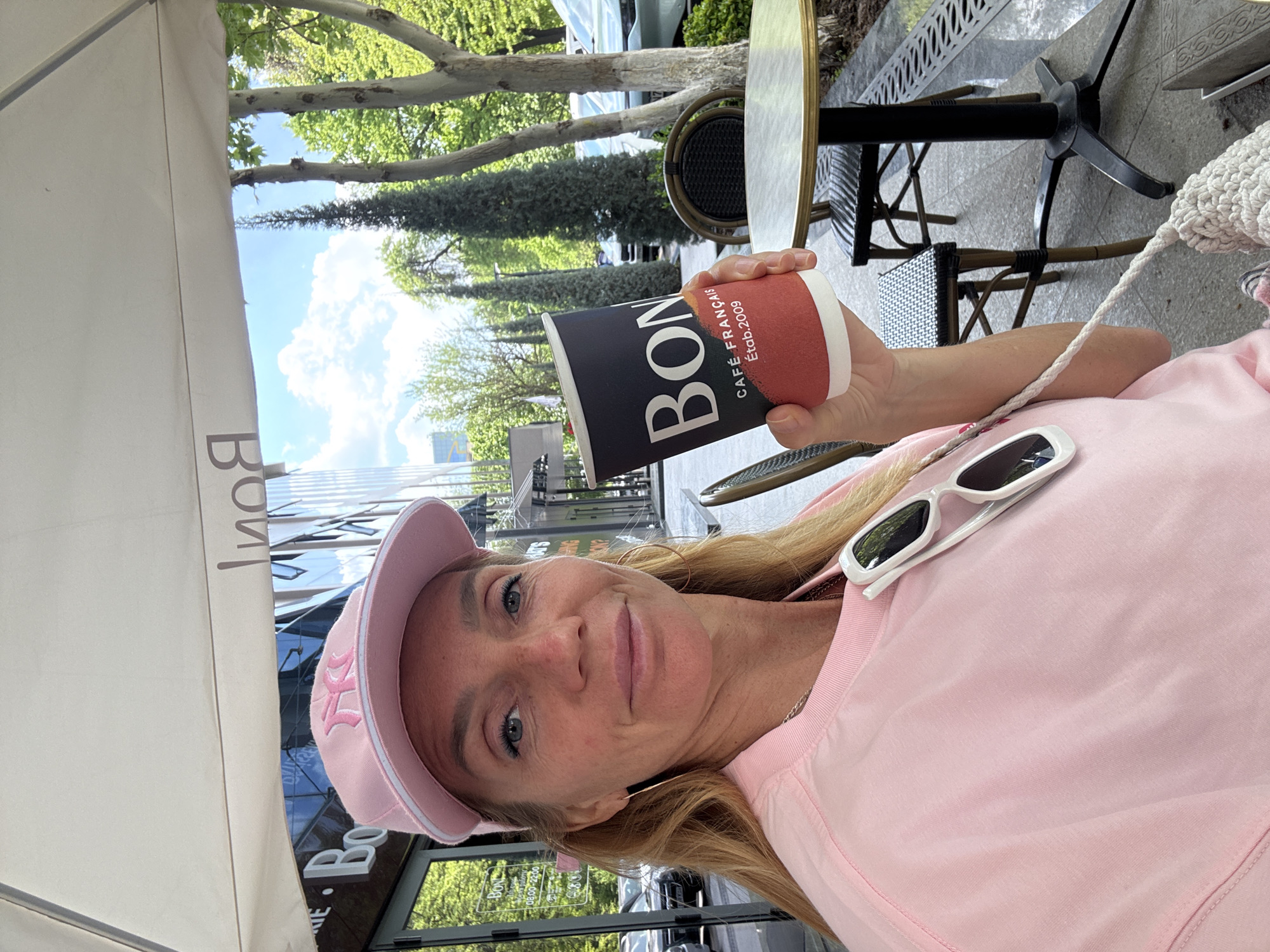 A woman is pictured in a selfie at an outdoor cafe in Tashkent, Uzbekistan. She's wearing a pink New York Yankees baseball cap and a pink sweatshirt, with white sunglasses resting on the fabric. She is holding a "BON!" coffee cup with text indicating it's a French cafe, established in 2009. The cafe has black bistro-style furniture. Overhead, a tan umbrella provides shade, and in the background, there are trees and a modern glass building.