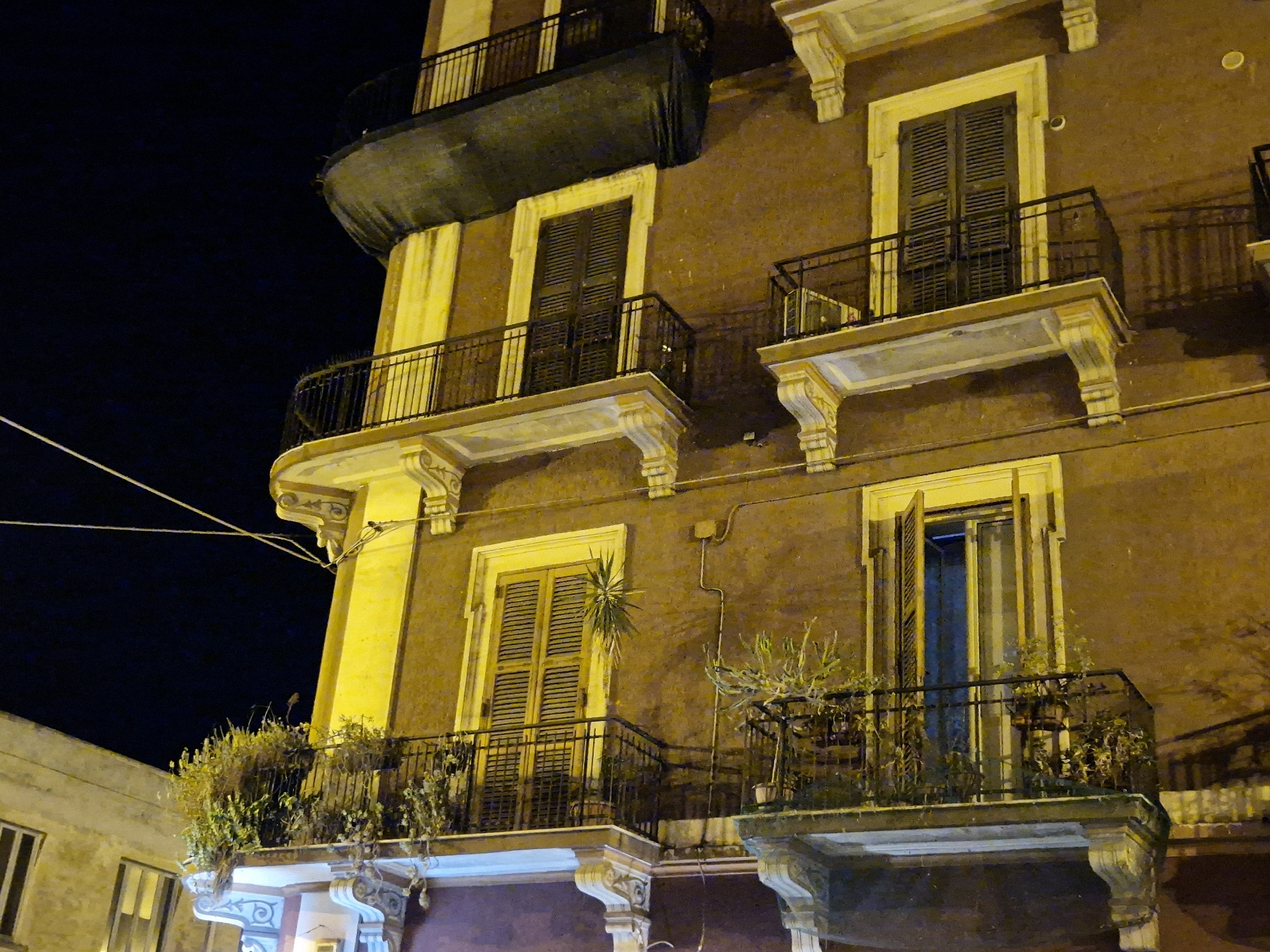 The image captures a nighttime view of a multi-story building in Taranto, Italy, with ornate balconies and windows illuminated by warm, artificial light. The building's facade is a warm brown color, with yellow trim around the windows and along the edges of the balconies. Several balconies are visible, each with black wrought-iron railings. Some balconies feature plants, adding a touch of greenery to the scene. The windows have dark shutters, some of which are closed, while others appear to be partially open, revealing glimpses of the interior. The sky is a deep, dark blue, indicating it is nighttime. The overall impression is one of historical architecture bathed in a warm glow against the night sky.