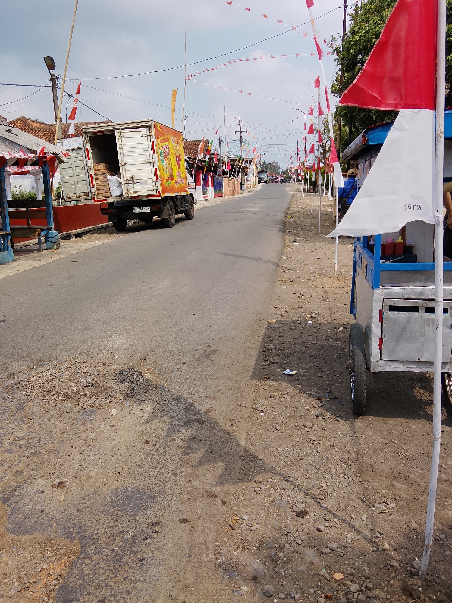 The image depicts a street scene adorned with Indonesian flags and pennants, suggesting a national celebration. In the foreground, a food cart with a blue trim and a white canopy stands on the right side of the road. The cart contains condiments such as ketchup and chili sauce. Further down the street on the left, a delivery truck is parked, its cargo bay open, revealing stacked goods. The truck has a colorful advertisement on its side. Numerous small flags, predominantly red and white, are strung along the street, attached to poles and wires. The road is paved but appears somewhat worn, with patches of gravel and debris. Buildings with red-tiled roofs line the street, and trees are visible in the background. The sky is cloudy. The overall impression is of a lively, festive atmosphere in a somewhat rural or suburban setting.