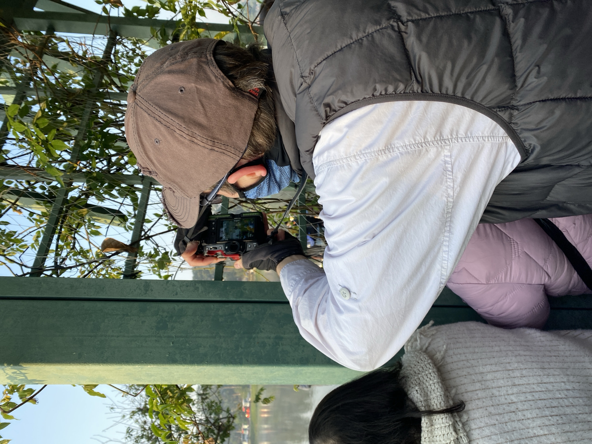 The image captures a moment on a bridge overlooking Xuan Huong Lake in Da Lat, Vietnam. A person wearing a brown baseball cap, black vest over a light-colored long-sleeved shirt, and black gloves is holding a digital camera and taking a picture. They are positioned behind a green metal railing, with climbing plants growing along it. Another person wearing a light-colored sweater and a pink jacket is partially visible in the lower part of the frame. The background shows the lake with vegetation and possibly buildings on the far side.