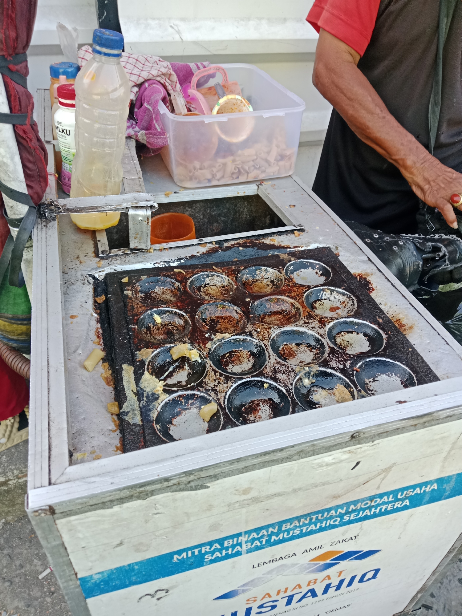 A street vendor is preparing snacks on a hot plate. The vendor, wearing a red shirt and black apron, is out of focus. The hot plate has multiple circular molds for cooking the snacks. Several snacks are in different stages of cooking, with some appearing partially cooked and others finished. There are also some cut pieces of food scattered around the hot plate. In the background, there are plastic bottles of drinks, a pink cloth, and a clear plastic container holding more food items. The vendor's cart has text on its side, suggesting it is part of a business support program. The scene appears to be outdoors, likely on a street in Kediri City, Indonesia, indicated by the text on the cart.