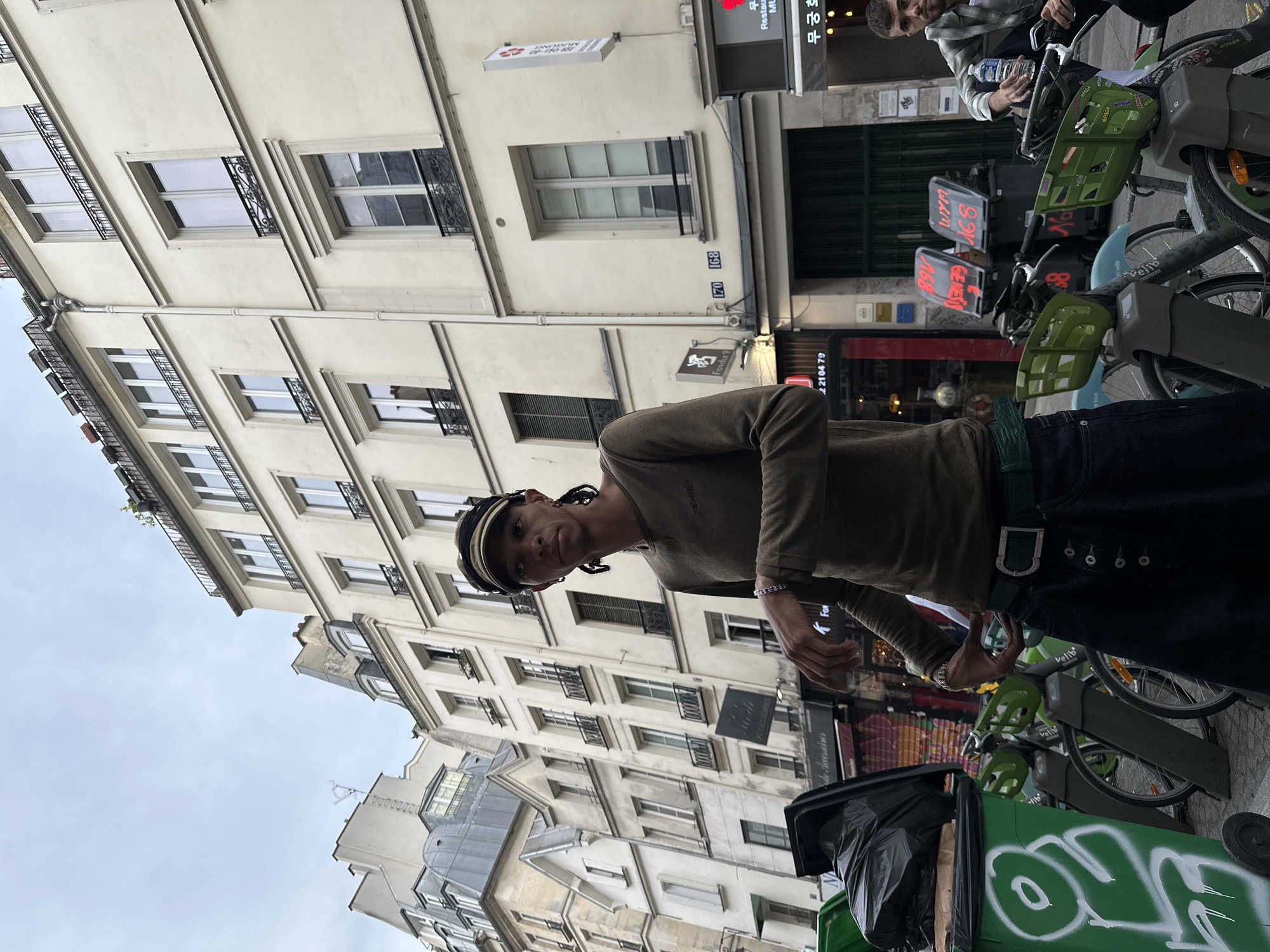 The image depicts a man on a street in Paris, France, standing near a Vélib' bike-sharing station and a trash receptacle. The man has dark skin, long dreadlocks, a light-colored cap, and is wearing a light brown long-sleeved shirt and dark jeans with a distinctive belt. He is holding something in his hand.
Behind the man, there are rows of Vélib' bikes. Another individual, partially visible, is by one of the bikes.
The background shows typical Parisian architecture, with multi-story buildings featuring ornate windows and detailing. Signage indicates local businesses, including a Korean restaurant. The street is marked with numbers 170 and 168.