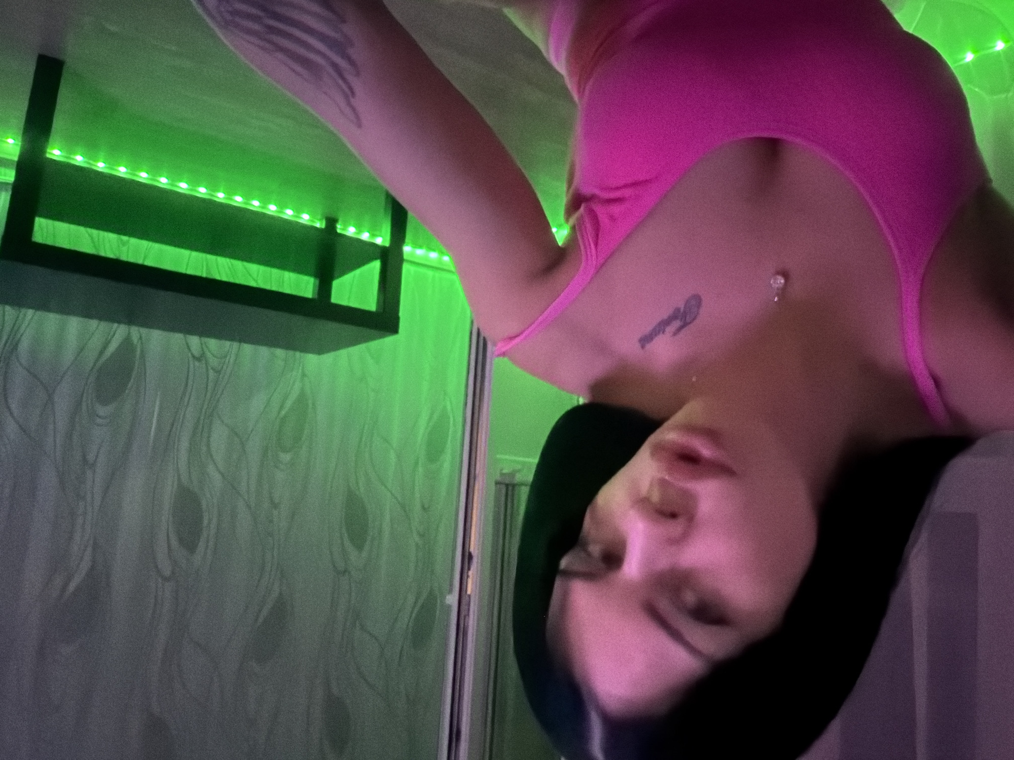 The media file depicts a young woman in a pink outfit, lying down with her head slightly tilted. Her face is turned towards the camera, with her eyes open and a neutral expression. She has dark hair and visible tattoos on her arm and chest. A belly button piercing is also visible. The background is illuminated by bright green LED lights, which create a neon glow. A dark shelf is mounted on the wall above her. The overall setting appears to be an indoor space, possibly a bedroom or living area, with patterned wallpaper visible. The lighting and the woman's pose suggest a casual or relaxed moment. There is no discernible time of day or weather information. The location context provided is Ufa, Russia, but there are no specific visual cues to confirm this. No text is visible in the image.