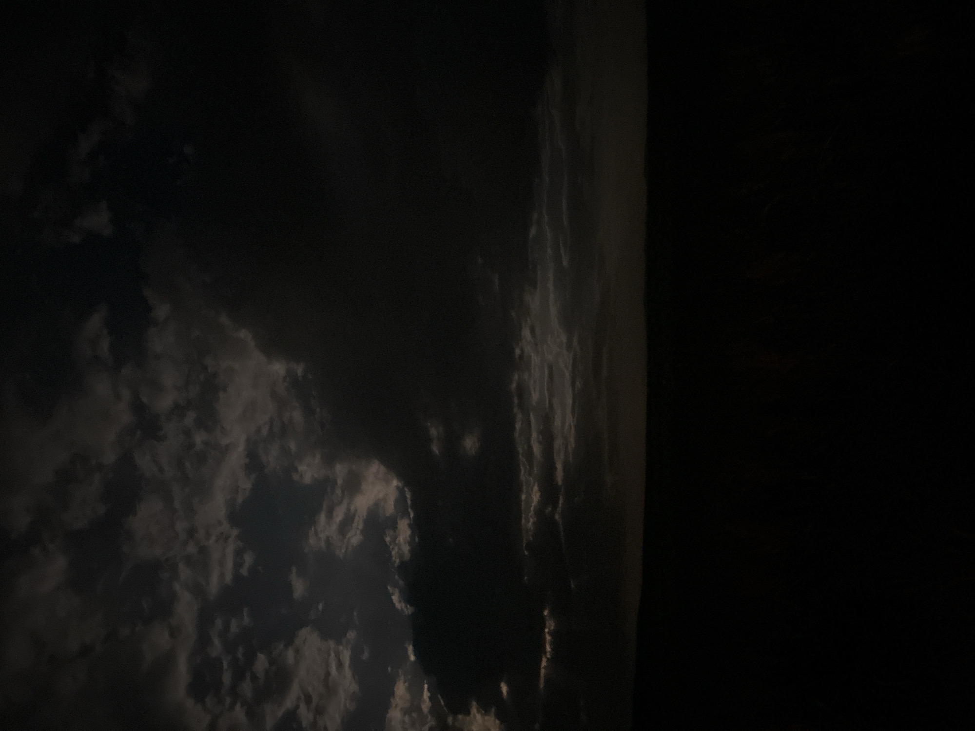 The image shows a dark, partially cloudy night scene. The left portion of the image is filled with scattered clouds, faintly illuminated. The right portion is completely dark, obscuring details. The image is not clear enough to determine the location or any actions, subjects, or interactions.