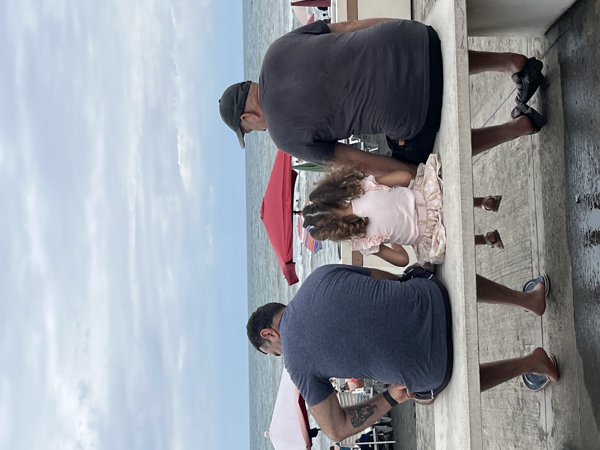 Three individuals, two adult males and a young girl, are seated on a concrete bench facing a beach and the Black Sea in Ureki, Georgia. The man on the left is wearing a dark blue t-shirt and shorts, with a tattoo on his left arm and a black watch on his wrist. He is barefoot with blue and white sandals. The girl in the center is wearing a light pink top with ruffled sleeves and a floral skirt. She has her hair in pigtails and is wearing sandals. The man on the right is wearing a dark gray t-shirt and black shorts, and a dark baseball cap. He is wearing dark sandals. The beach scene behind them includes beach umbrellas, lounge chairs, and other beachgoers. The sky is overcast with a light gray cloud cover. The sea is calm with small waves.