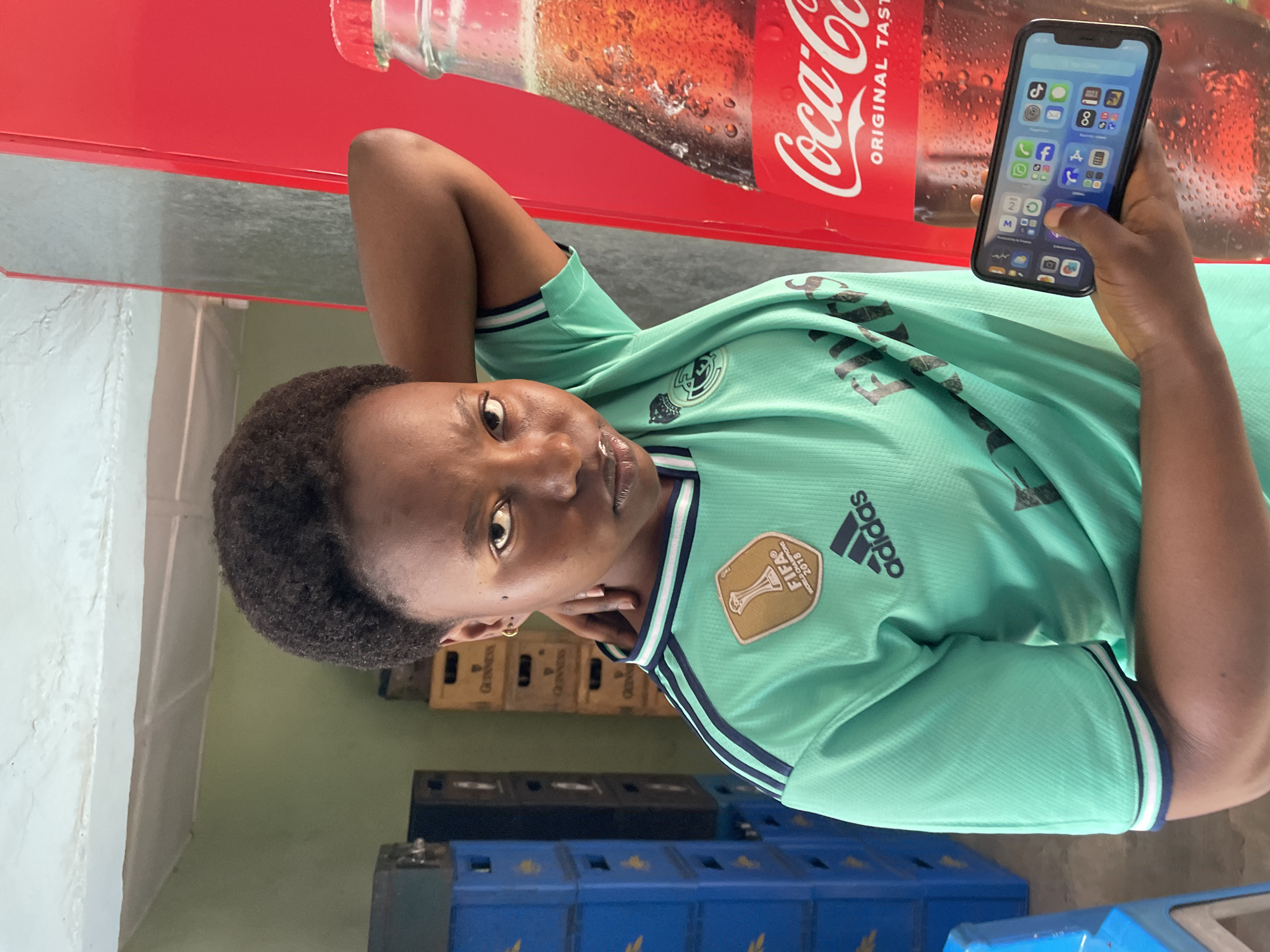 The image captures a medium shot of a young woman in an indoor setting. She is wearing a light green t-shirt with dark blue stripes on the collar and sleeves, and a FIFA World Champions 2018 badge on the left chest, along with the Adidas logo. Her right arm is bent, with her hand resting on the back of her neck. In her left hand, she holds a smartphone displaying various app icons. The background shows stacks of blue plastic crates on the left and wooden crates, some with Guinness branding, in the middle. A large red Coca-Cola refrigerator with a visible bottle of Coca-Cola is on the right. The lighting suggests an indoor environment, possibly a shop or storage area. The overall scene is likely within a commercial area in Jos, Nigeria, given the context provided.