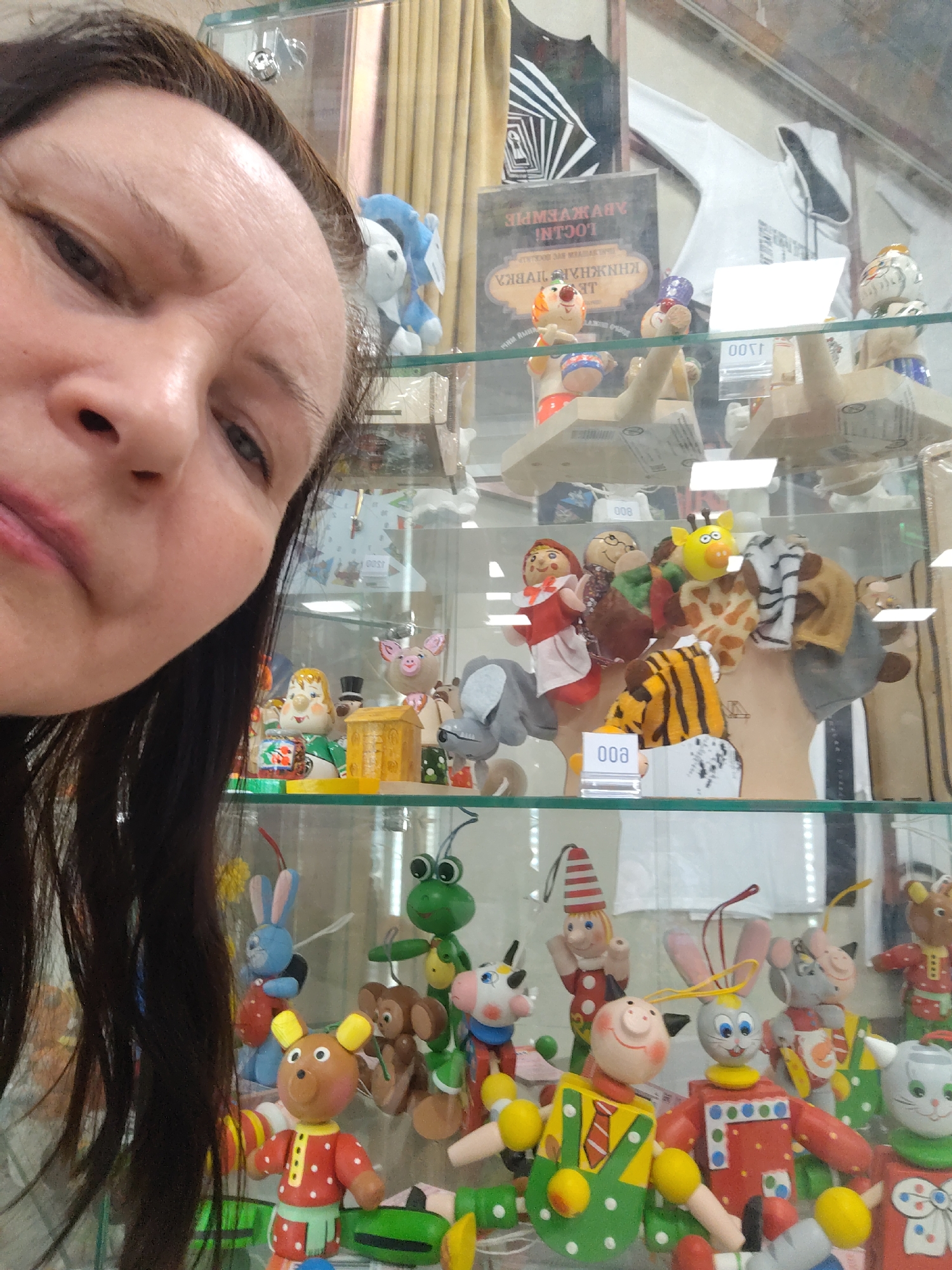 A woman is taking a selfie in front of a glass display case filled with various wooden toys and puppets. The toys include figures of animals like bears, rabbits, frogs, and pigs, as well as human characters. The puppets on the middle shelf appear to be finger puppets of animals such as giraffes and tigers, as well as characters like Little Red Riding Hood. In the background, behind the display case, there are other items for sale, including clothing and a book with Cyrillic text. Based on the Cyrillic text and style of the merchandise, the location is likely a shop or market in Moscow, Russia.