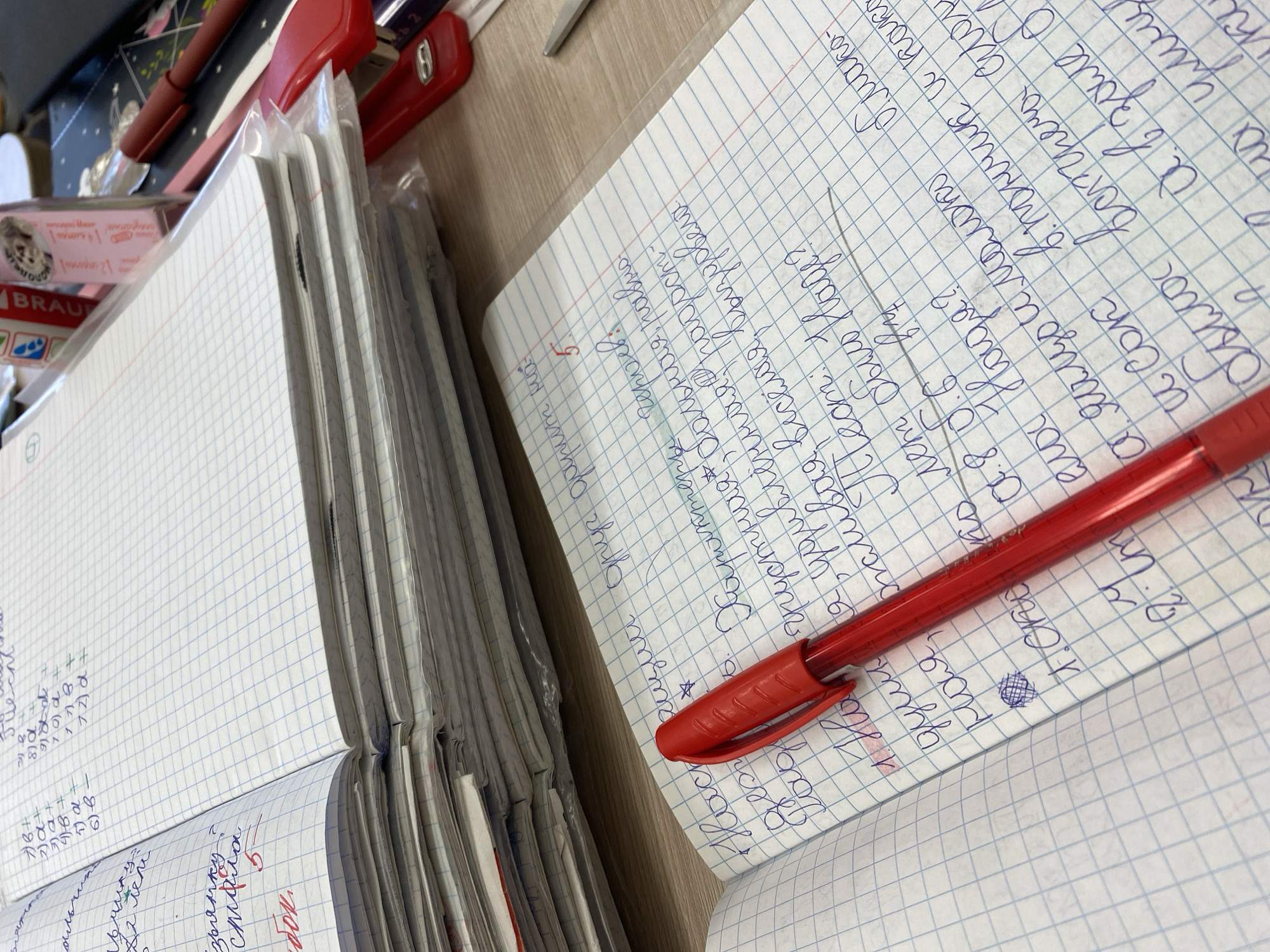 The image shows an open notebook filled with handwritten text in blue ink, next to a stack of similar notebooks. The open notebook also has a red pen resting on it. The writing appears to be in Russian. There is another open notebook partially visible at the left edge of the image. A red stapler, as well as an unopened box of Braun ear thermometers are partially visible in the background. The setting appears to be a desk or table. The location might be Chaikovsky, Russia as some of the text includes proper names written in Russian.