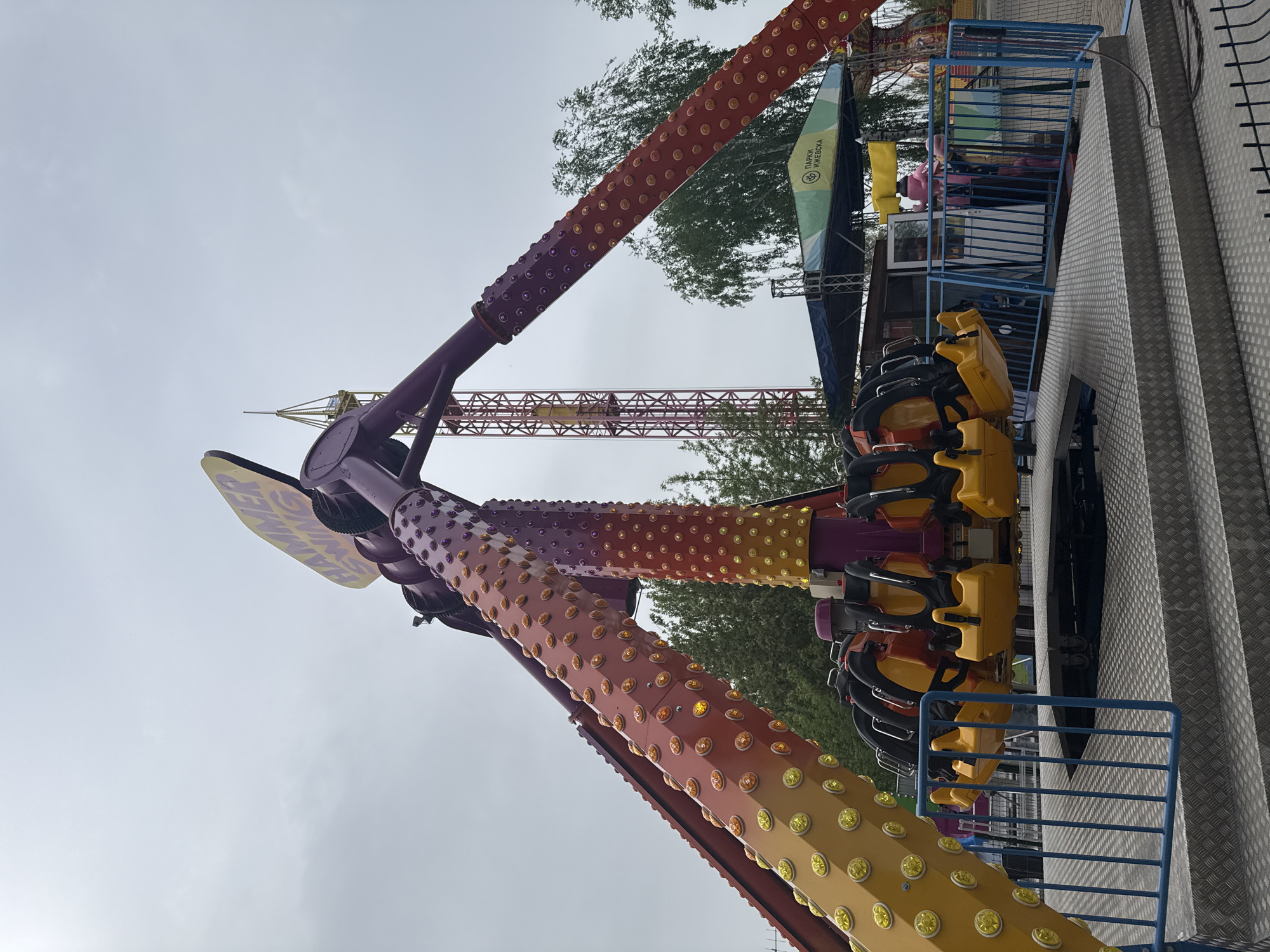 The image captures a portion of a vibrant amusement park ride in Izhevsk, Russia, known as "Flaming Hammer." The ride features two long arms in an orange, yellow, and purple color scheme adorned with circular, light-up decorations. Seating modules with bright yellow seats are attached to the end of one arm. The ride is set against an overcast sky, and in the background, a tall, red-and-yellow drop tower can be seen, along with green trees. The perspective is slightly angled upward, making the ride appear towering. A metal safety platform with a blue railing is visible on the right side. A sign in Russian, identifying the location as "Парки Ижевска" (Parks of Izhevsk) is partially visible near the ride's control booth. People can be seen inside the control booth.