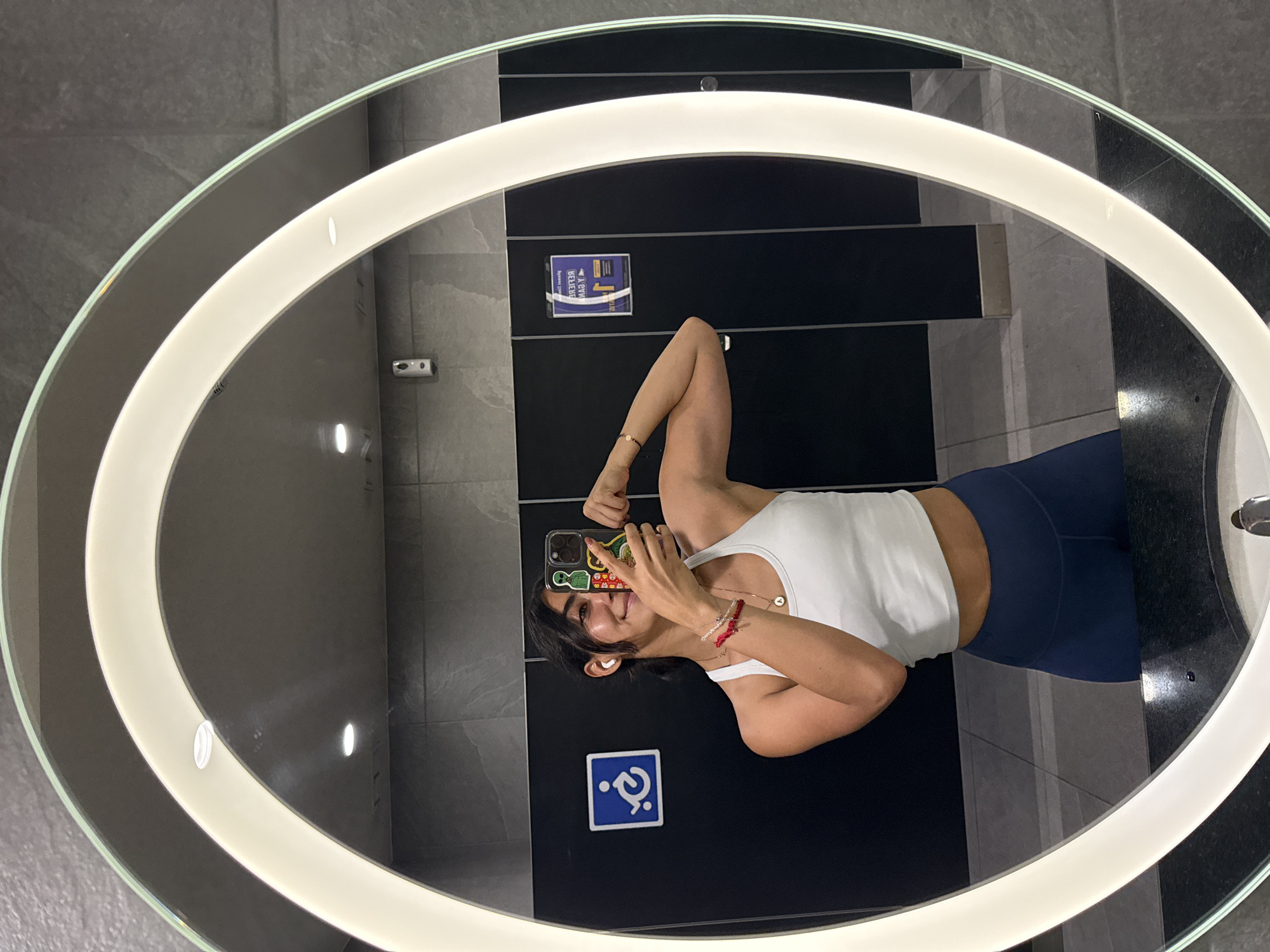 The image shows a woman in a restroom taking a selfie in a mirror. The mirror is oval-shaped and surrounded by a ring of light. The woman is wearing a white tank top and blue leggings. She is holding her phone up in front of her face. The restroom has gray and black tiles. A sign with the international symbol of access is visible on the wall. There is also a sign with Spanish writing on the wall. The location of the scene is Puebla, Mexico.
