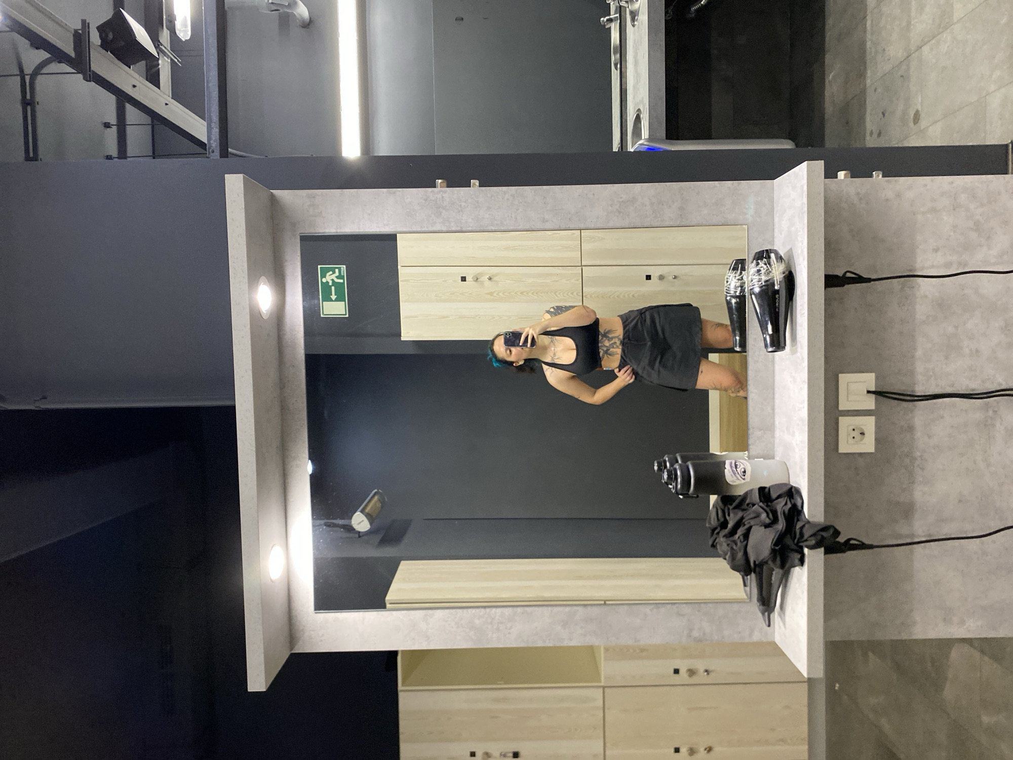 A woman is photographing herself in a mirror. The mirror is in a bathroom or dressing room with gray walls and wooden lockers. There are hair dryers and electrical outlets mounted on the wall to the right of the mirror. There is a green and white exit sign to the left of the mirror. The location is most likely in Gijón/Xixón, Spain, but not confirmed.