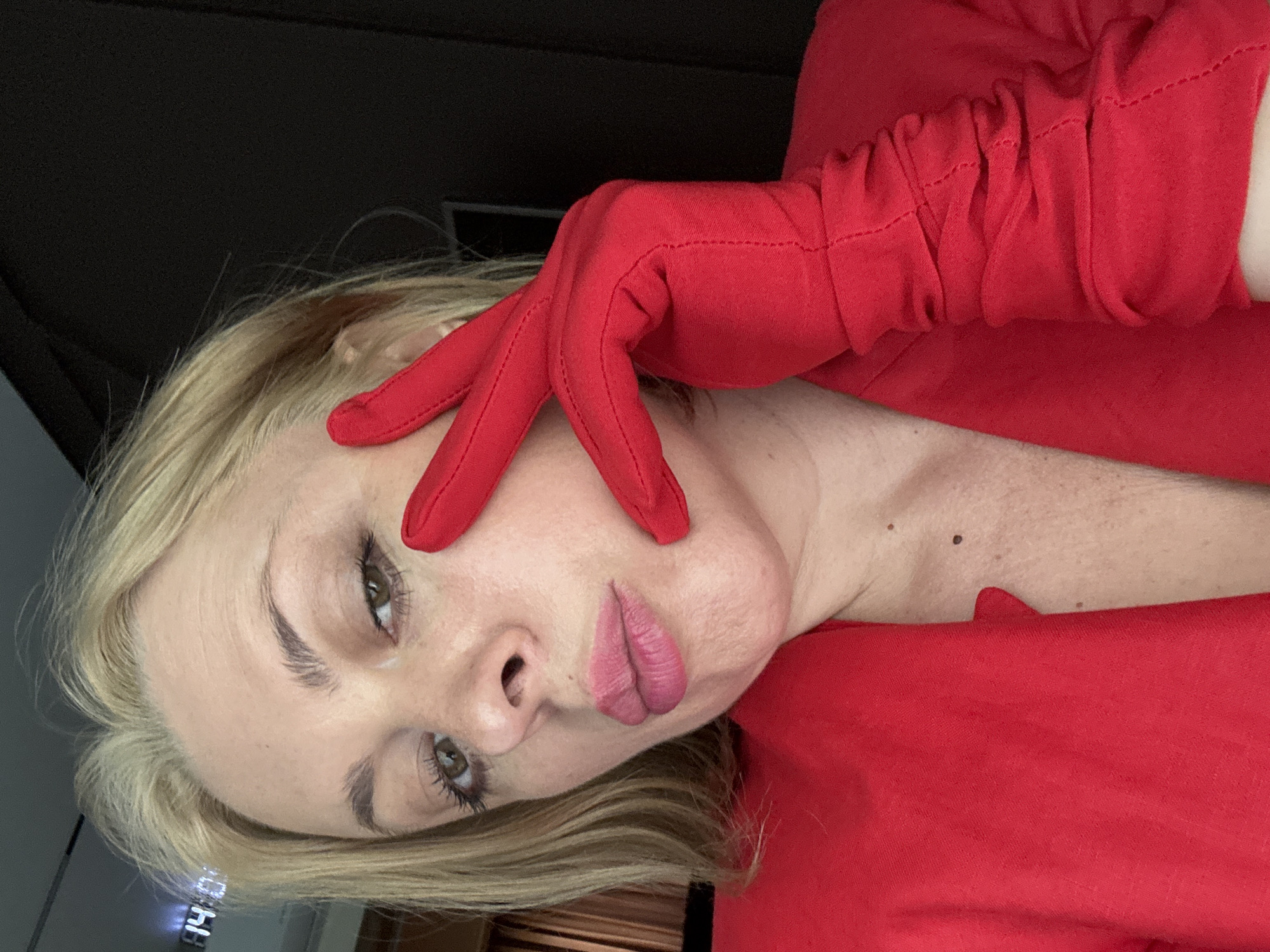 The image shows a close-up of a woman's face and upper body, who is wearing a red blazer and red gloves. Her right hand is raised to her face, with her fingers touching her cheek and the side of her nose. She has blonde hair, brown eyes, and is wearing pink lipstick. The background appears to be indoors, with a dark wall and a faint digital clock displaying "14:08" on the left side of the image. The scene does not contain any specific landmarks or features that would identify it as being in Sochi, Russia.