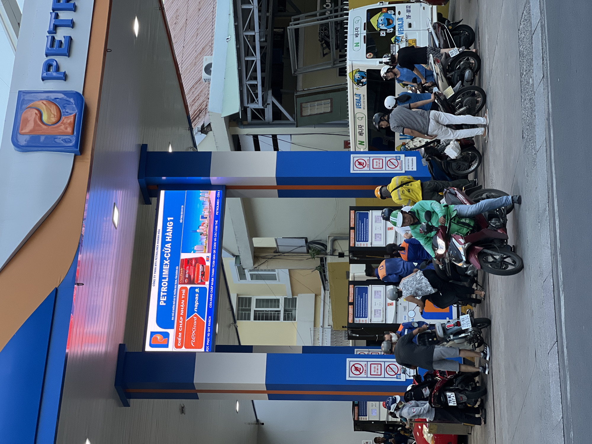 The image shows a busy gas station in Nha Trang, Vietnam, with several people on motorcycles and a white van. The gas station is branded with "PETROLIMEX" and features a large digital display screen. People are seen in casual attire, many wearing helmets, suggesting they are either refueling their motorcycles or waiting in line. The environment appears to be a typical urban setting with buildings and a road in the background.