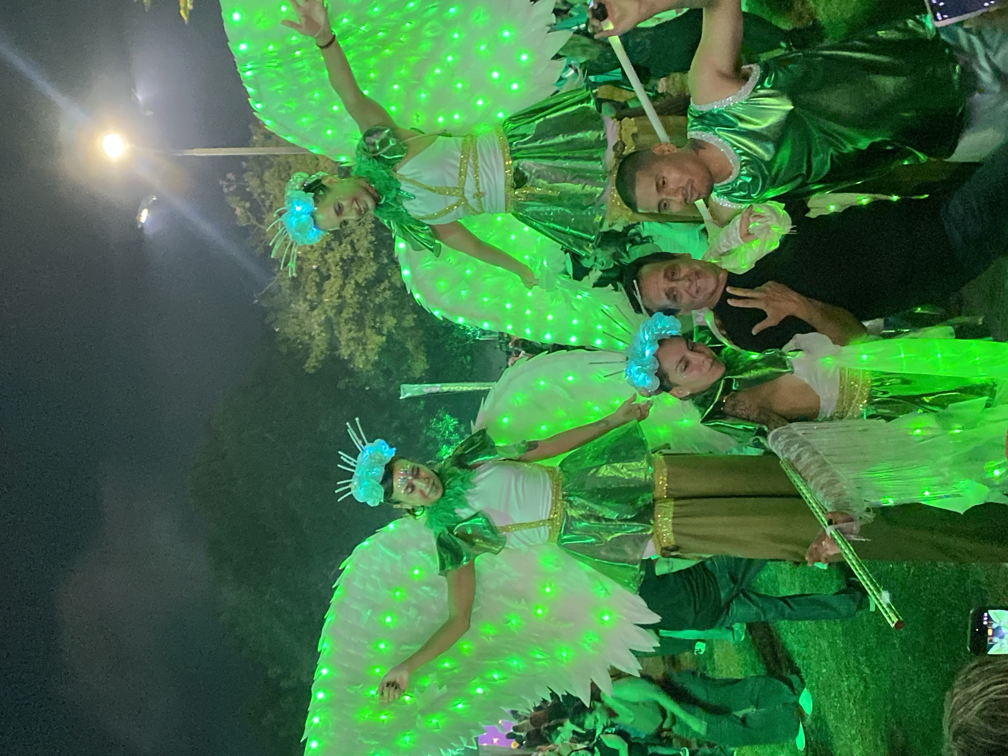 The image shows a group of people at night. Two women wear white and green costumes with glowing wings and floral headpieces. Two men in green and black shirts stand near them. One man makes a peace sign. The setting appears to be outdoors, possibly a parade or festival in Buenos Aires, Argentina, given the vibrant costumes and public nature of the gathering. A streetlight is visible in the background. People are holding phones in the scene.