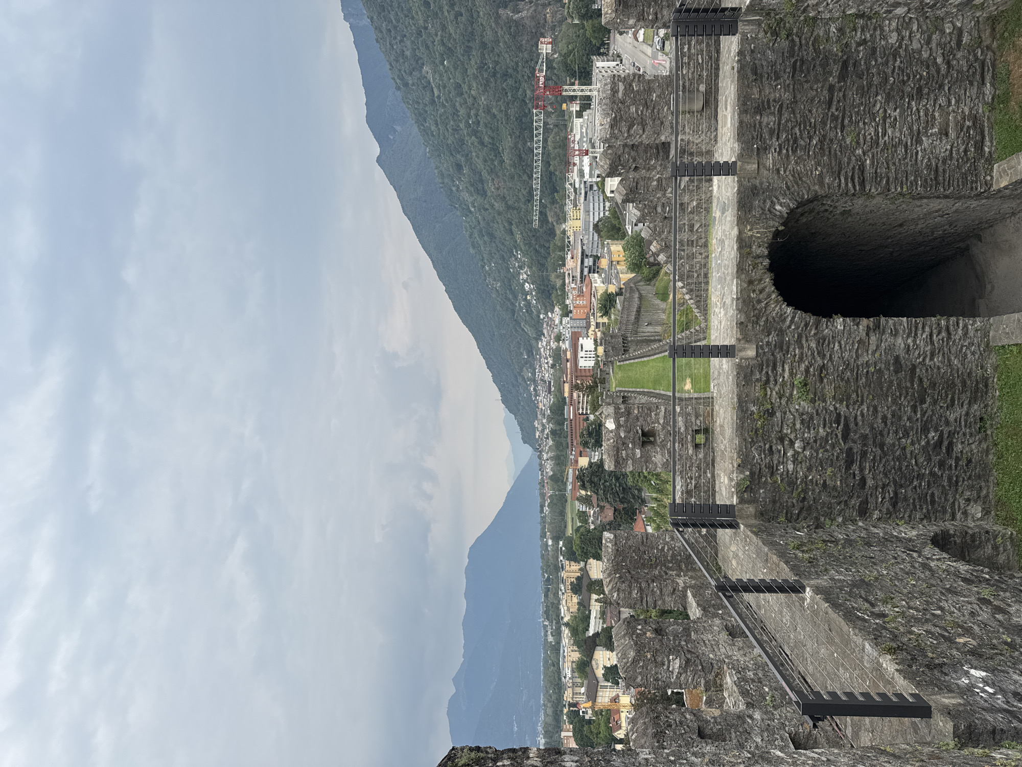 This image captures a view from the battlements of Castel Grande in Bellinzona, Switzerland. The foreground is dominated by the stone walls and walkways of the castle, featuring a dark arched entryway leading down stone steps. A metal railing with wire mesh runs along the walkway. Beyond the castle walls, a green grassy area stretches out, leading to a long, straight pathway bordered by grass. Further in the distance, the city of Bellinzona unfolds with a collection of buildings, some modern and some older, interspersed with trees. Two prominent red and white construction cranes are visible within the cityscape, indicating ongoing development. The city is nestled at the base of steep, verdant mountains that dominate the background. The sky is overcast with heavy, grey clouds.