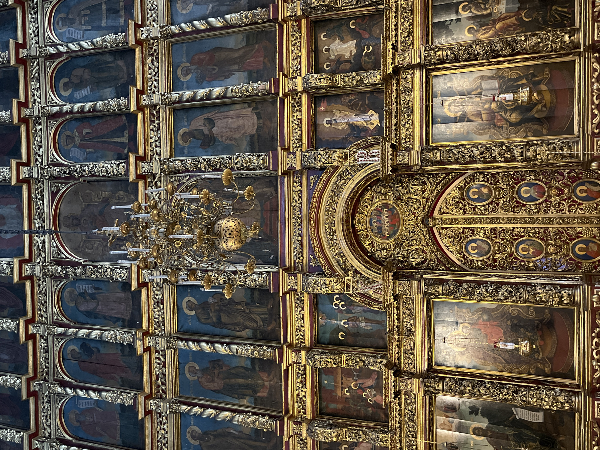 The image depicts the interior of the Cathedral of the Nativity of the Virgin Mary in Pskov, Russia. The scene is dominated by a large iconostasis, a wall of religious icons, elaborately gilded and adorned with intricate carvings. The iconostasis is divided into multiple tiers and rows, each filled with framed paintings of saints and biblical scenes. A grand chandelier hangs from the ceiling, casting a warm glow on the opulent display. The icons themselves are richly detailed, with figures rendered in traditional Byzantine style. The overall impression is one of immense historical and spiritual significance, showcasing the artistic and religious heritage of the Orthodox Church.