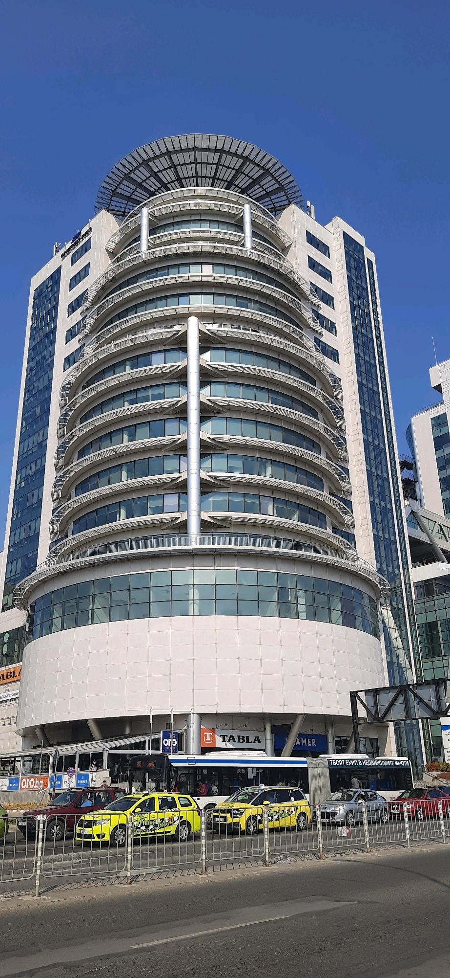 In Varna, Bulgaria, a modern, circular skyscraper with a helipad on top stands against a clear blue sky. The building's facade is a mix of white panels and large glass windows, with distinctive curved balconies on the upper floors. At the base of the skyscraper, businesses are visible, including "TABLA" and "AMER". In the foreground, a street scene unfolds with several vehicles: two yellow taxis, a dark red SUV, and a silver car, all lined up behind a metal barrier. A bus is also present in the traffic. The overall impression is one of a bustling urban environment in good weather.