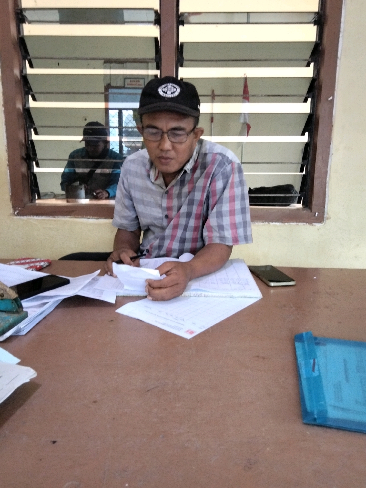 A man wearing a black cap and glasses sits at a brown desk in a room with a window. He is wearing a plaid shirt and is holding papers and a pen, appearing to be working. Through the window, another person can be seen reflected, and a Indonesian flag is visible hanging. The desk is cluttered with papers, a stapler, and cell phones. A blue folder is also on the desk. The environment appears to be an office or classroom setting. The image is taken from a medium shot, focusing on the man and the desk.