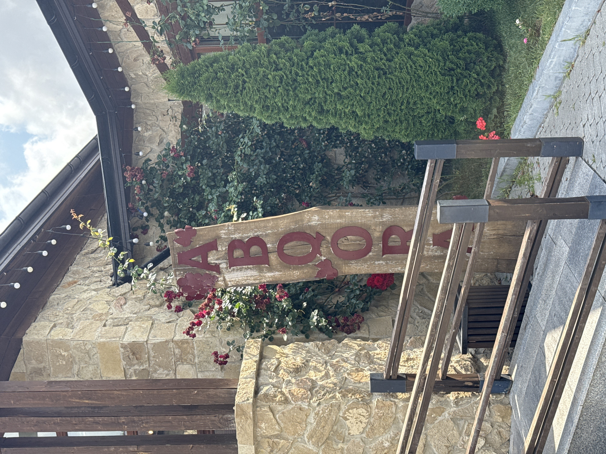 A weathered wooden sign with red lettering stands vertically near the entrance of a building. The letters spell out "AABOOR," with the last two letters partially obscured by foliage. The building's exterior is constructed of rough, light-colored stone blocks, and dark wooden beams are visible along the roofline and supporting the structure. Strings of small, round lights are strung along the overhang of the roof. Lush green foliage, including climbing plants and a tall, conical evergreen tree, surrounds the building. Red flowers bloom amongst the greenery. A wooden railing with horizontal planks leads up a ramp to the entrance. The ground is paved with grey stones and concrete. The scene appears to be an outdoor area of a establishment in Mirikənd, Azerbaijan.