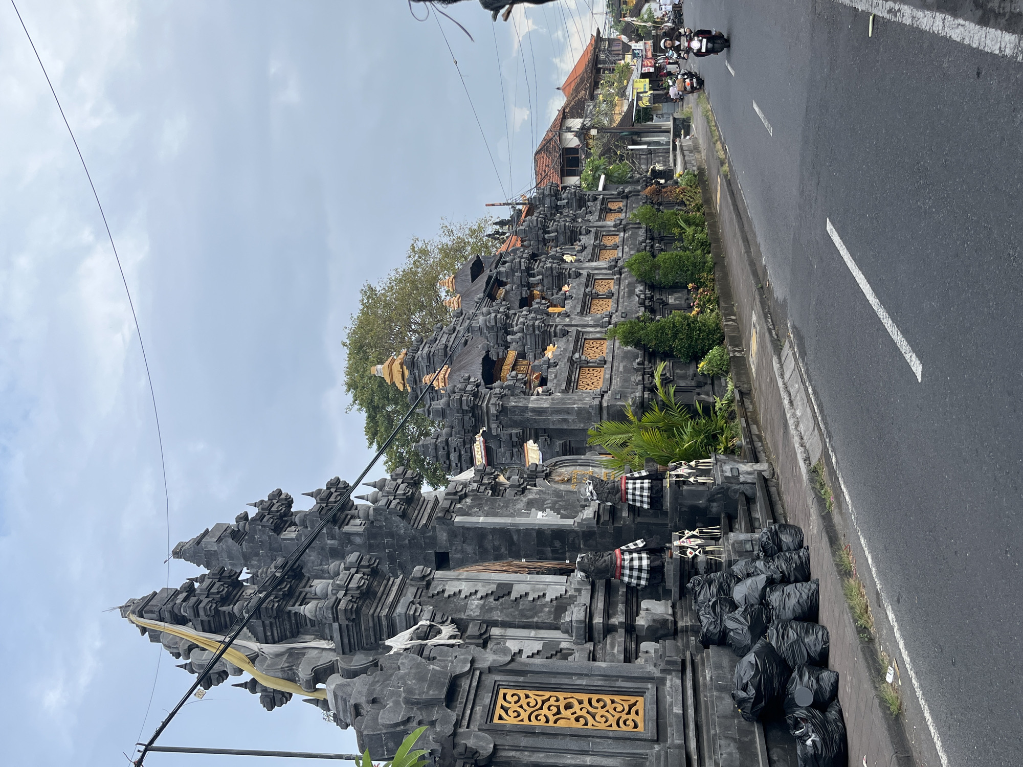 A street scene in Kuta Selatan, Indonesia, features a Balinese temple gate on the left, constructed of dark stone with intricate carvings and gold accents. A yellow and white cloth is draped over one of the gate's spires. In front of the gate, a row of black garbage bags is piled along the sidewalk. Two stone statues, dressed in black and white checkered cloths, stand guard at the entrance. Lush green plants and small trees line the temple wall. Across the street, motorbikes are visible on the asphalt road, with a few buildings in the background. The sky is overcast with a few streaks of light clouds.
