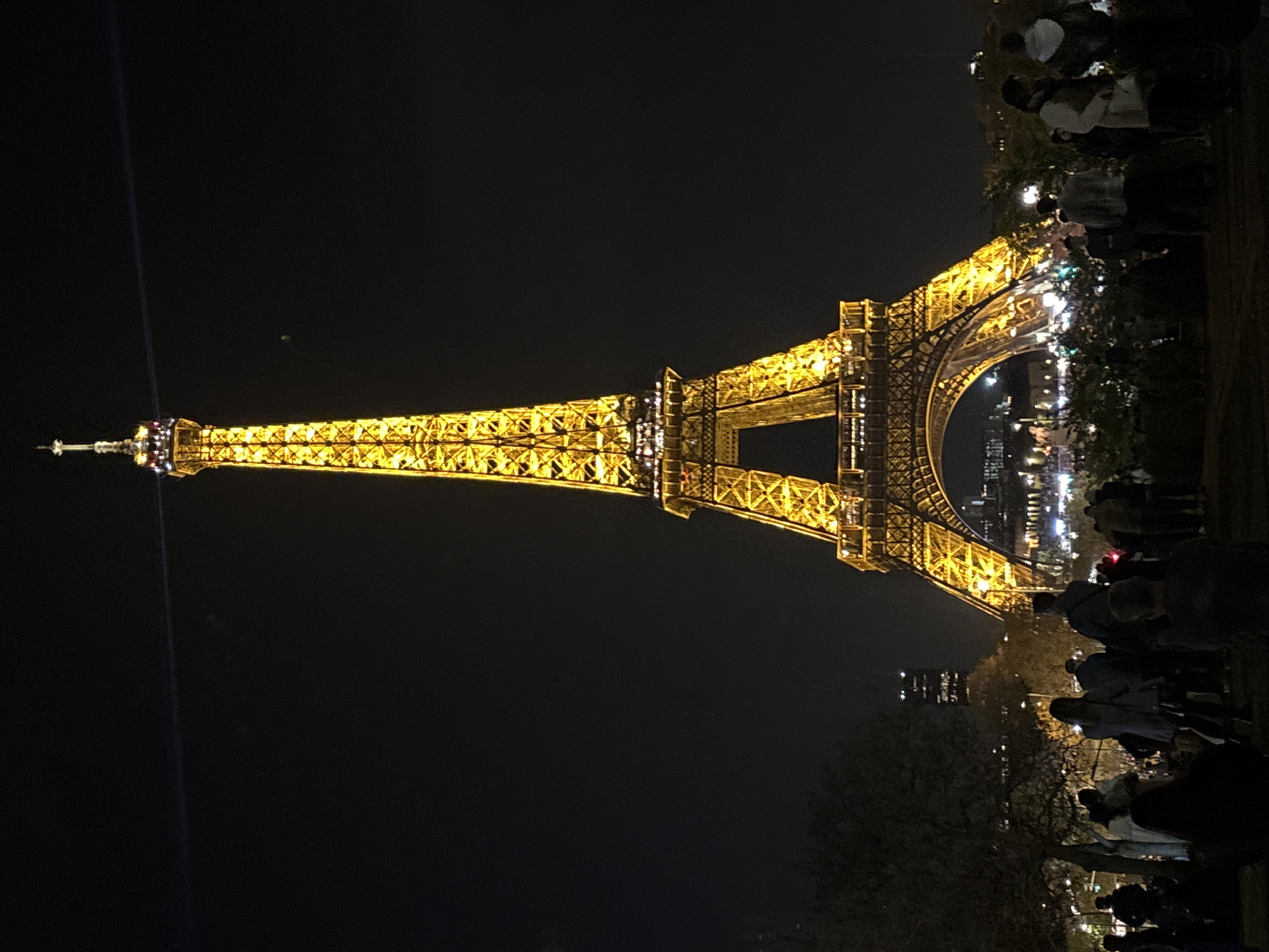 Illuminated at night, the Eiffel Tower dominates the view in Paris, France. Crowds of people are gathered near the tower. Buildings are visible in the background, and trees are also present near the tower. The night sky is dark.