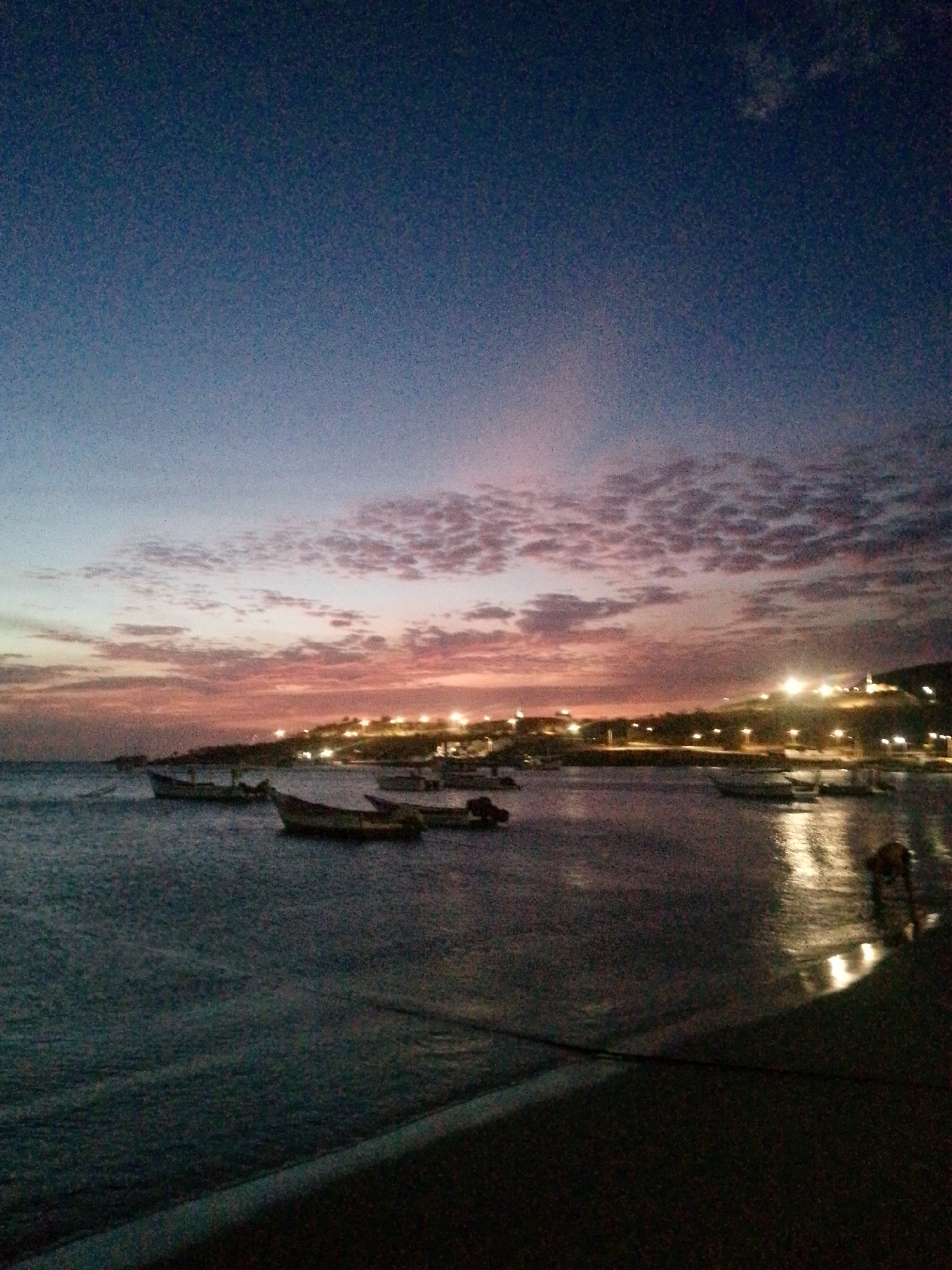 This is a photograph taken at dusk or twilight in Juan Griego, Venezuela. The image showcases a body of water, likely the ocean, with several small boats moored near the shore. The sky transitions from a dark blue at the top to a mix of orange, pink, and grey near the horizon, indicating a sunset. Above the horizon line are scattered clouds, also tinted with hues of the setting sun. In the distance, along the coastline, are lights that appear to be from buildings or street lamps, suggesting a populated area. The foreground is dark, obscuring detail, but appears to be a part of a structure or embankment next to the water. There are no visible people in the scene.